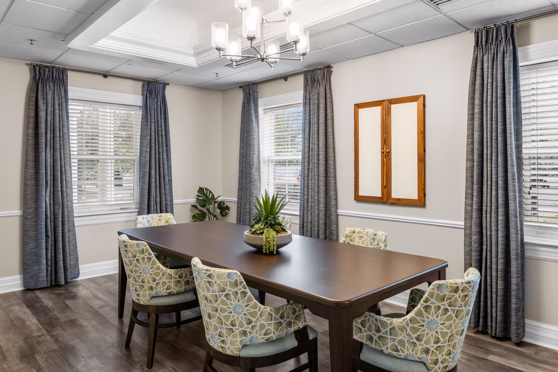 Bright dining room with a rectangular wooden table surrounded by six patterned chairs, a plant centerpiece, windows with gray curtains, and a chandelier.
