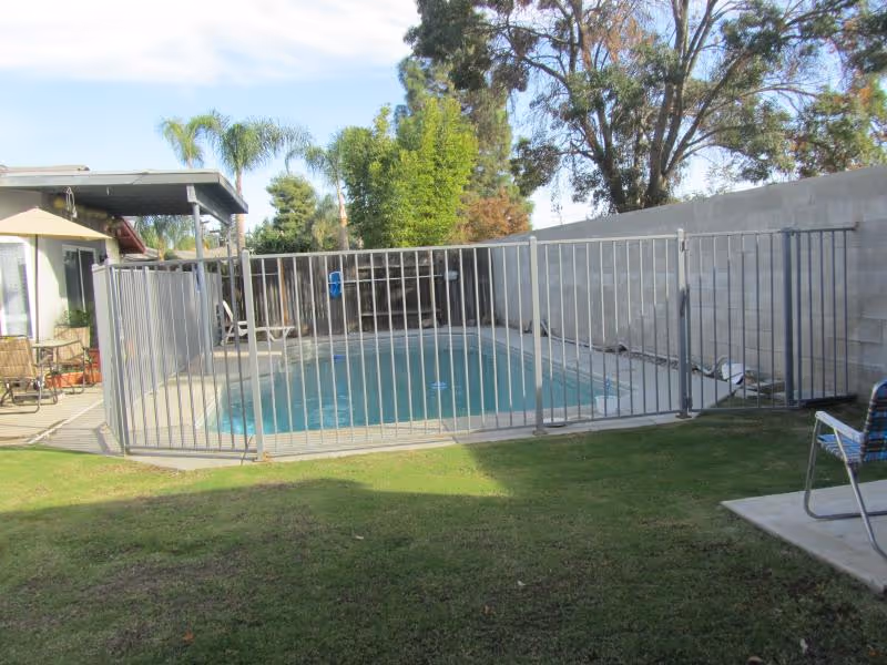 Backyard swimming pool enclosed by a metal safety fence with patio furniture and lawn.