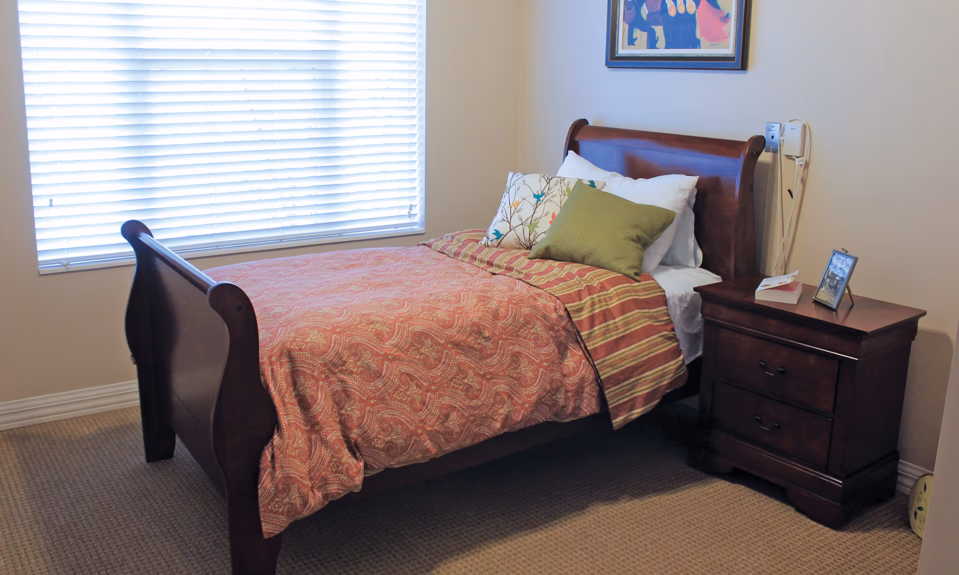 A bedroom with a wooden sleigh bed covered with a red patterned bedspread and striped blanket, decorated with three pillows including a green one. Next to the bed is a dark wooden nightstand with two drawers, a framed photo, and a book on top. A window with white blinds is on the left wall, and a colorful framed artwork hangs above the bed on the right wall.