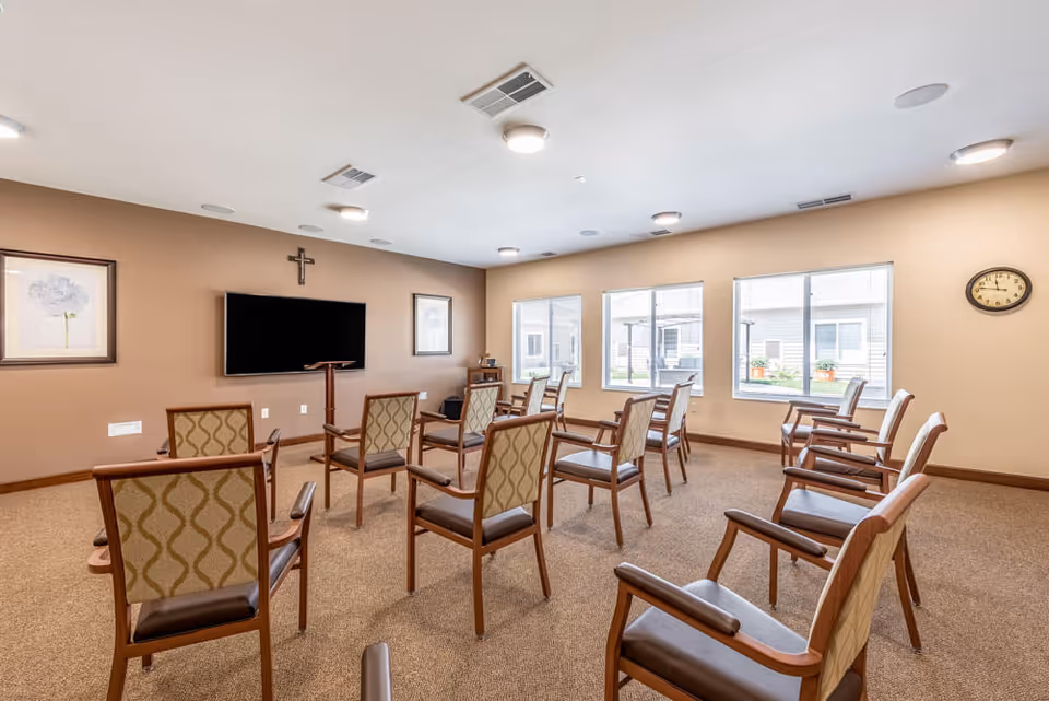 A well-lit room with beige walls and carpeted floor, arranged with multiple wooden chairs with patterned upholstery facing a wall-mounted flat screen TV and a wooden podium. The room has three large windows letting in natural light, two framed pictures on the wall, a clock, and a cross mounted above the TV.