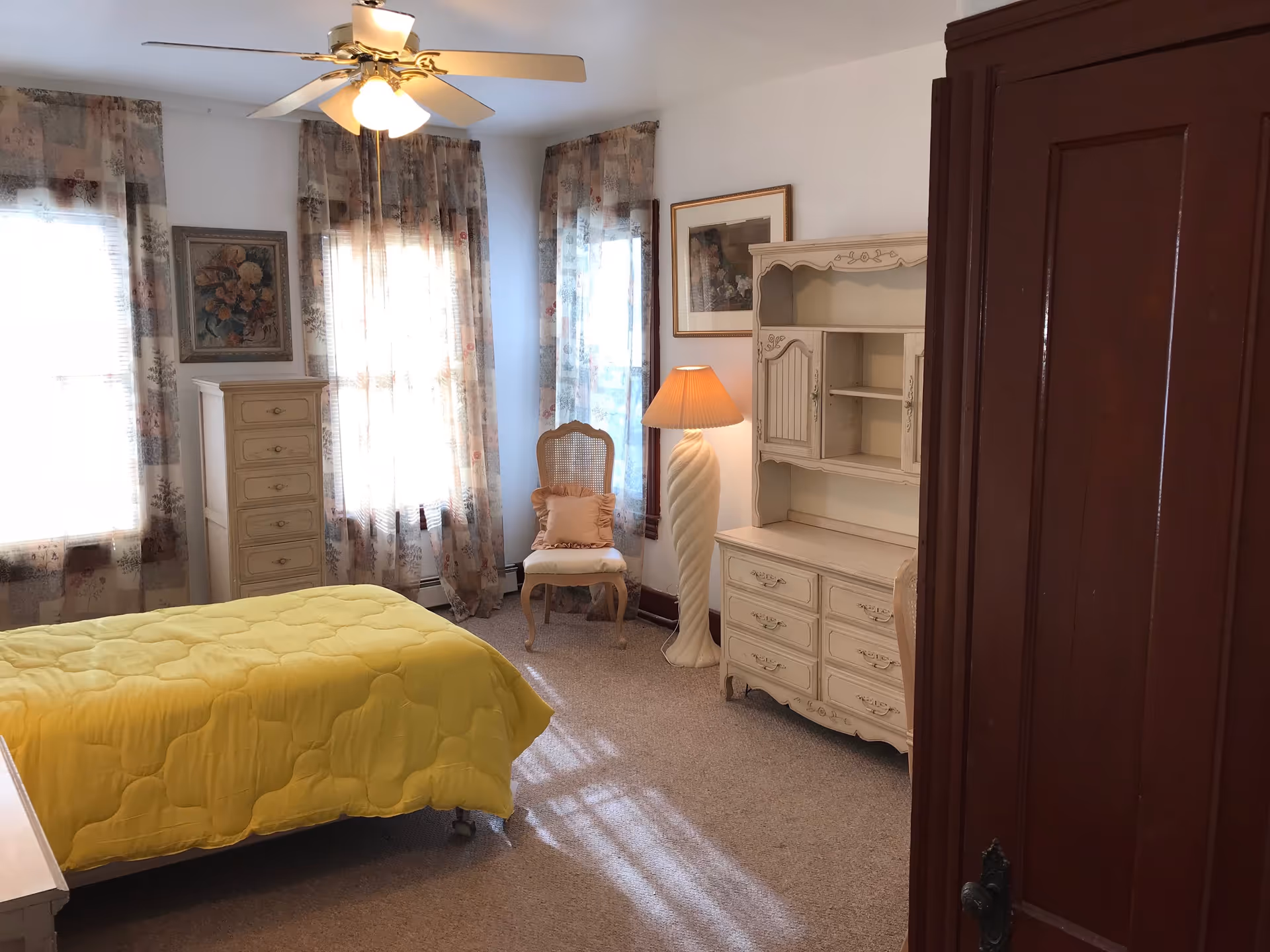 A cozy bedroom with a yellow bedspread on a single bed, floral curtains on three windows, a tall cream-colored dresser, a matching cream-colored hutch with drawers, a chair with a cushion, a standing lamp with a pleated shade, and framed artwork on the walls.