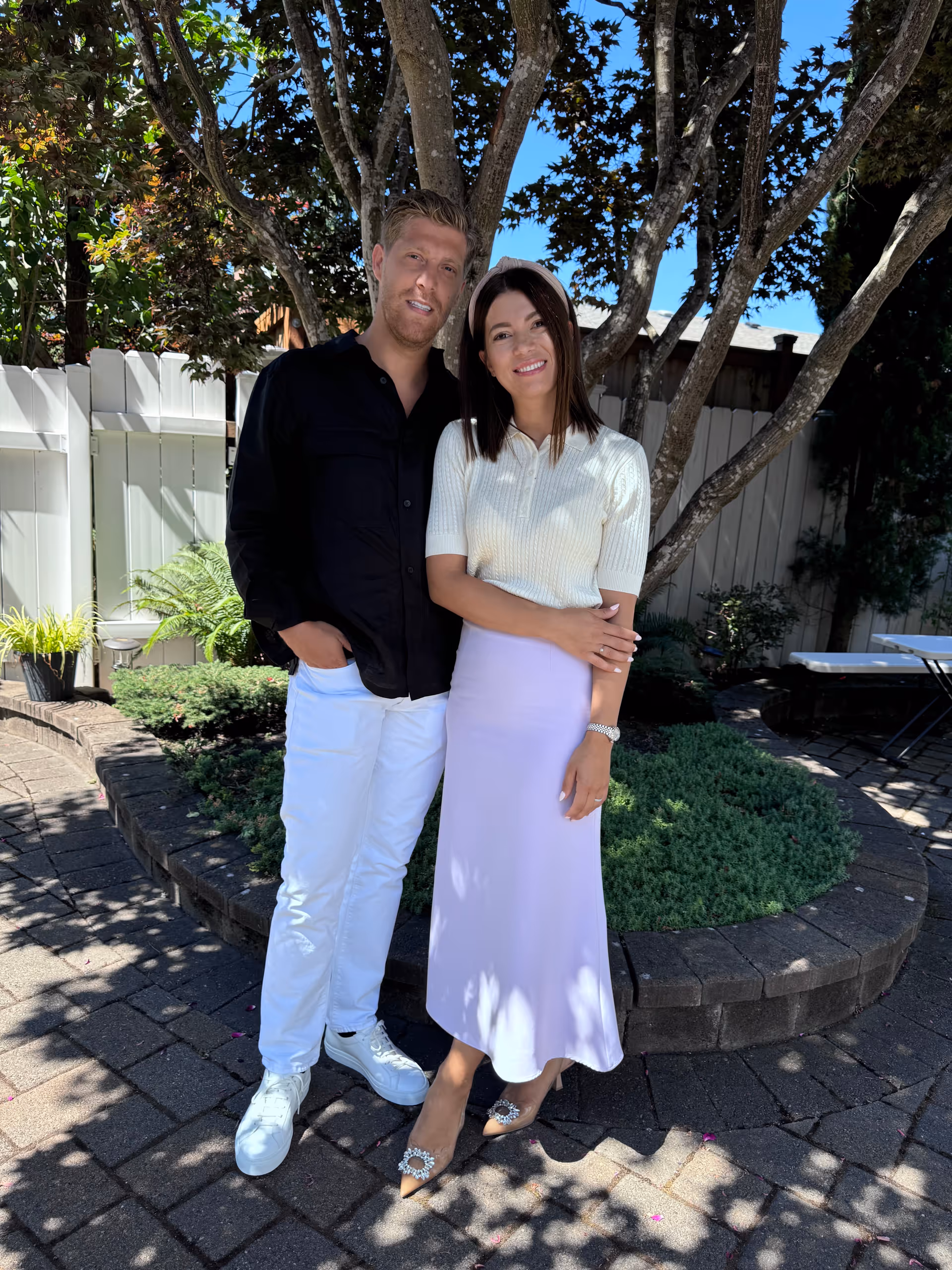 A man and a woman standing closely together outdoors on a paved area with a tree and greenery behind them. The man is wearing a black shirt and white pants with white sneakers, and the woman is wearing a white short-sleeve top and a light purple skirt with pointed shoes. They are smiling and posing for the photo in a garden-like setting with a white fence and some outdoor furniture visible in the background.