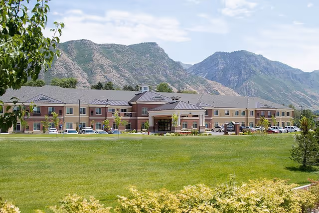 Front view of a large two-story senior living facility with a wide green lawn and mountains in the background.