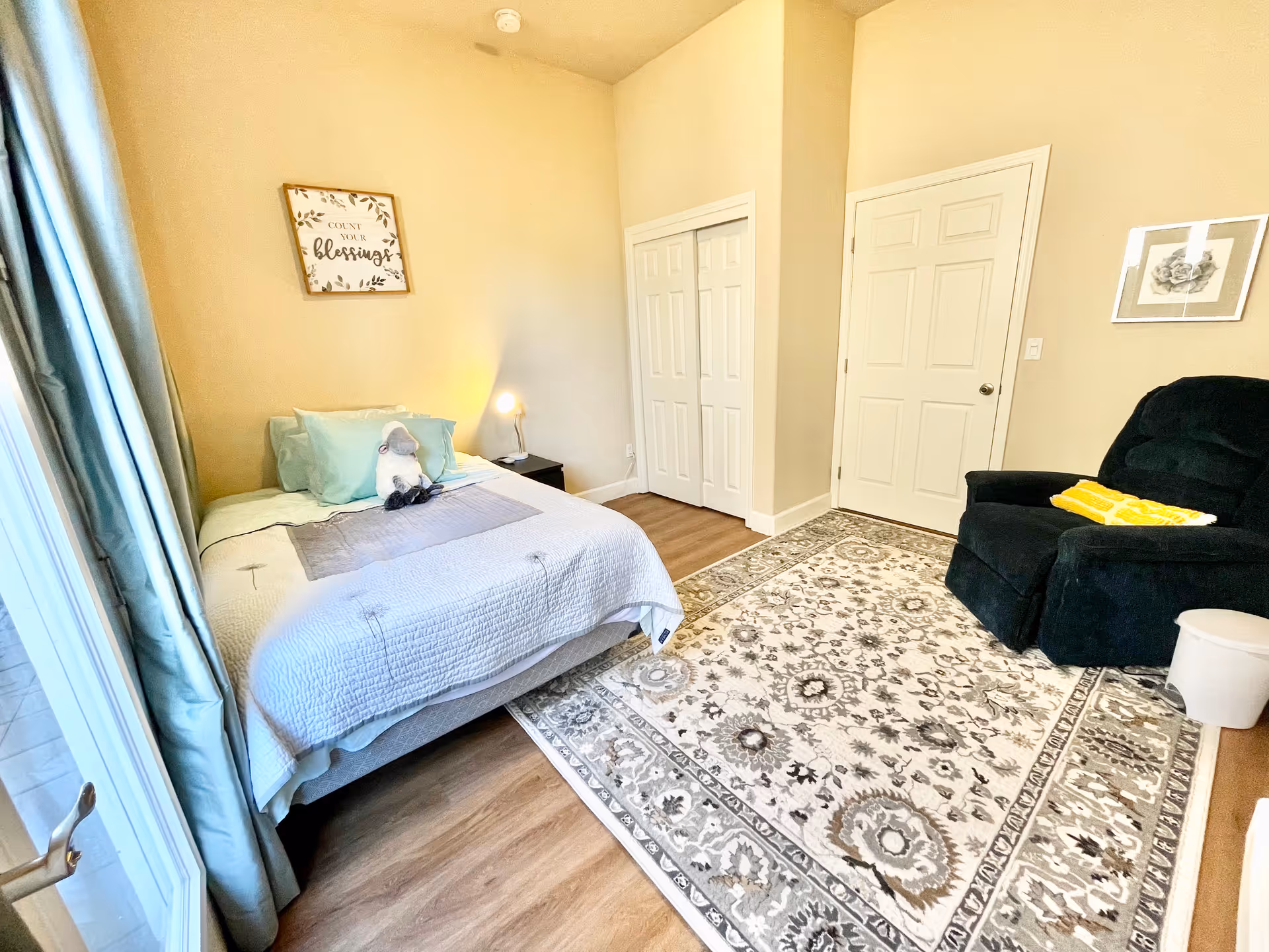 Sunlit bedroom with a bed, bedside table and lamp, a recliner chair, patterned area rug, and closet doors.