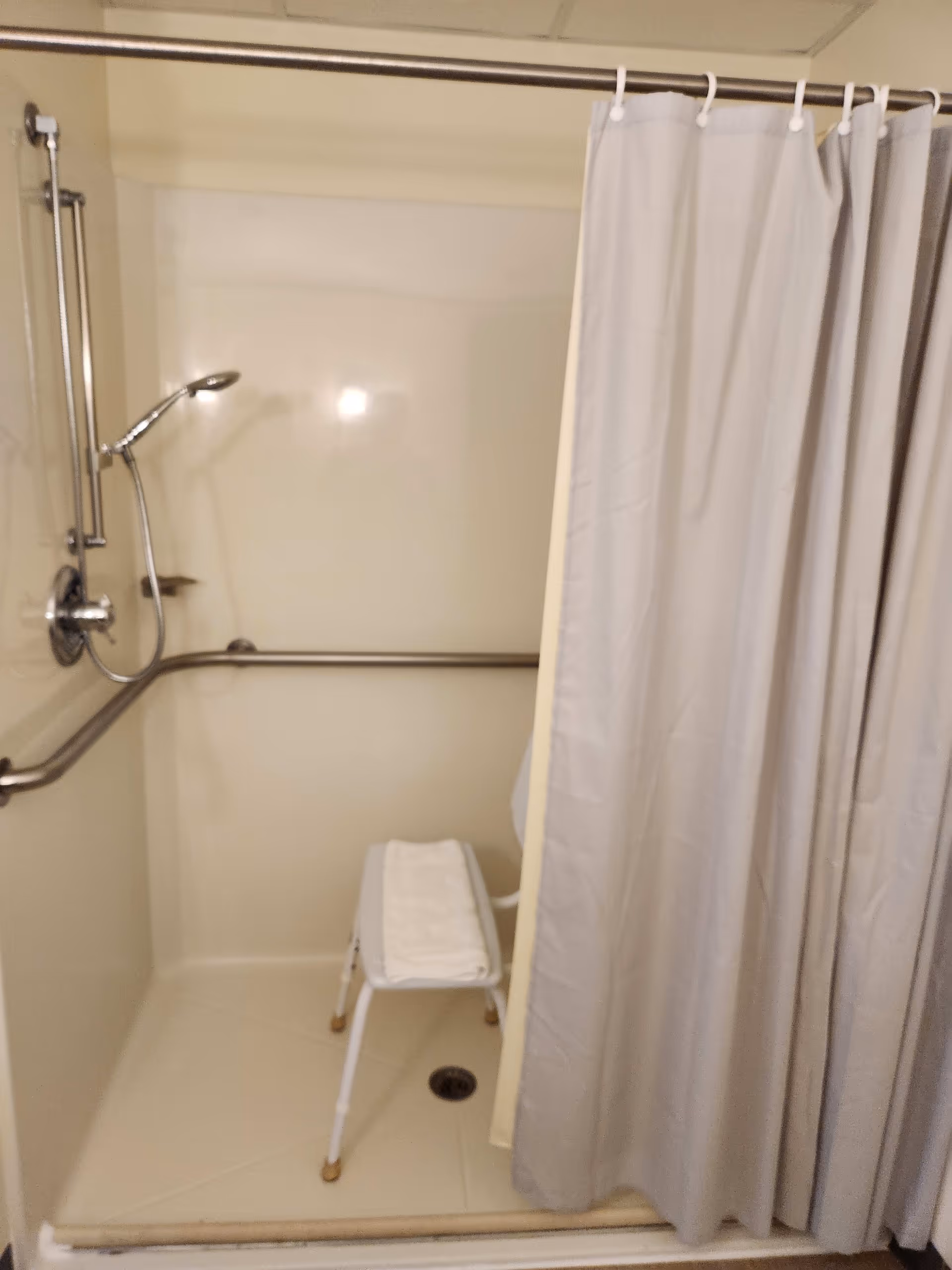 A shower area with a white shower curtain partially drawn to the side, a handheld showerhead mounted on a vertical bar, grab bars on the walls for support, and a white shower chair with a folded towel on it.