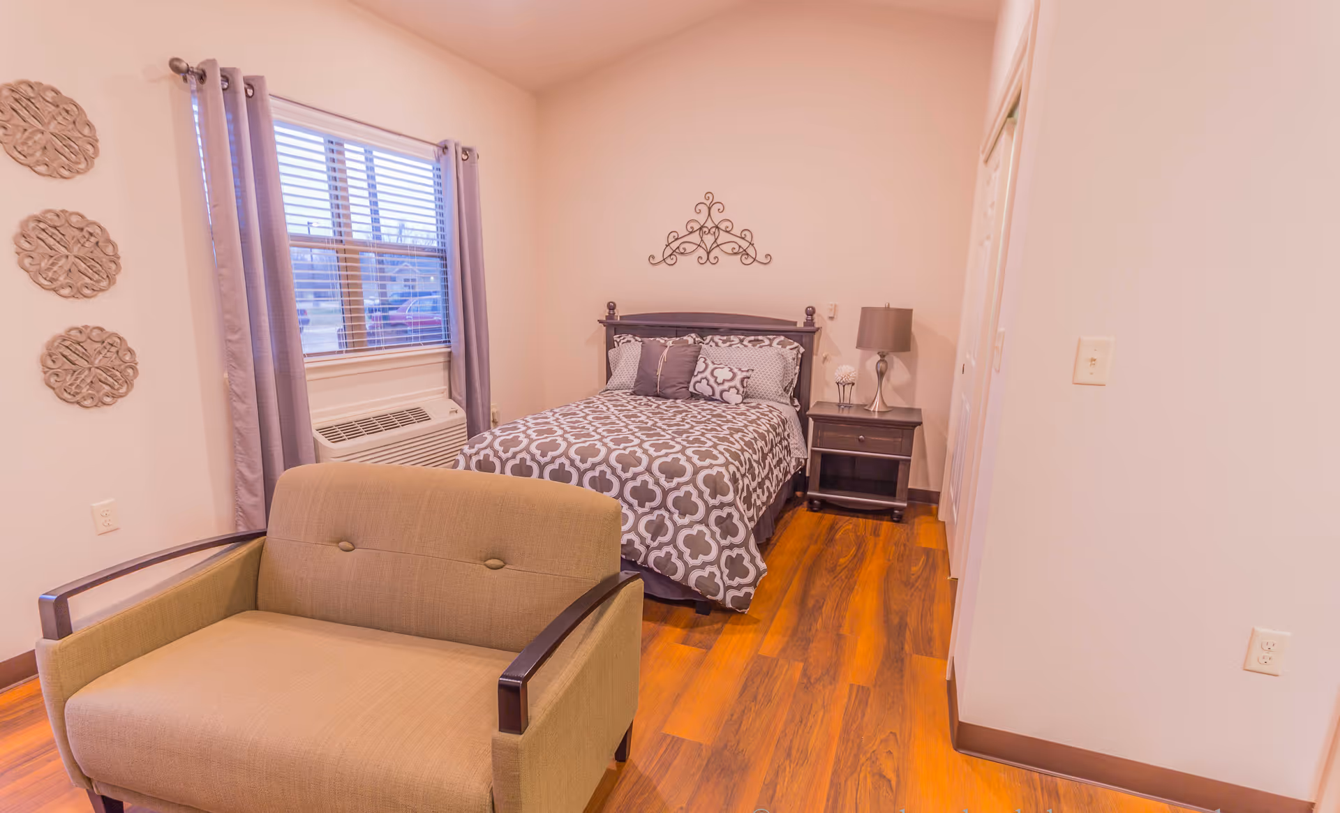 Furnished bedroom with a patterned bed, nightstand and lamp, a cushioned armchair, window with curtains, and wood-look flooring.