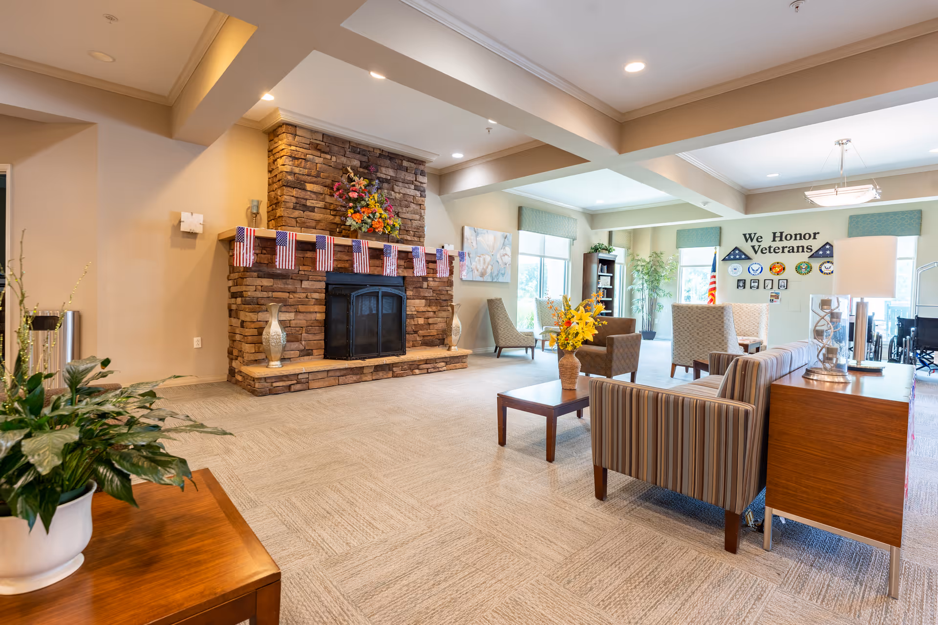 A spacious and well-lit living room area in a senior living facility with a stone fireplace decorated with small American flags and a colorful flower arrangement on the mantel. The room features comfortable seating including striped and patterned armchairs, a wooden coffee table with a vase of yellow flowers, and a side table with a lamp. Large windows allow natural light to fill the space, and a wall in the background displays a tribute to veterans with the text 'We Honor Veterans' and military emblems. There are also plants and bookshelves adding to the cozy atmosphere.