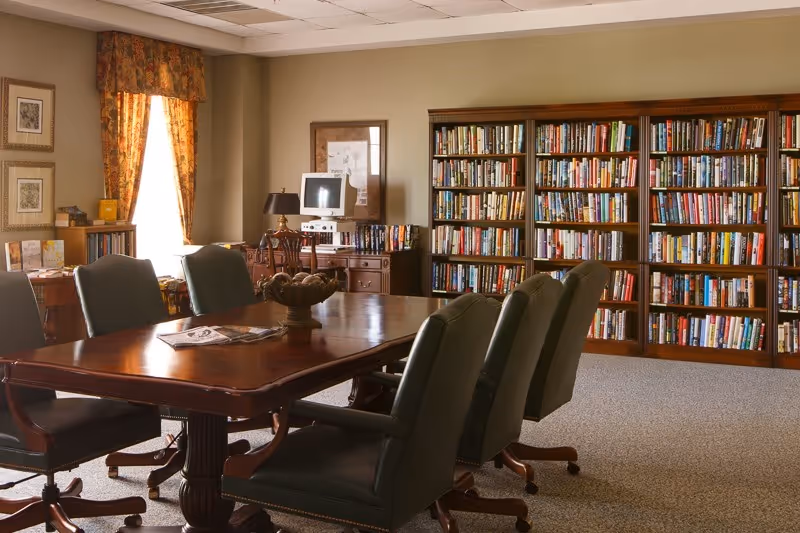 A cozy library or meeting room with a large wooden table surrounded by six green leather chairs on casters. Behind the table is a wall filled with bookshelves packed with books. There is a window with floral curtains letting in natural light, framed pictures on the walls, and a small desk with an old computer and lamp in the corner.