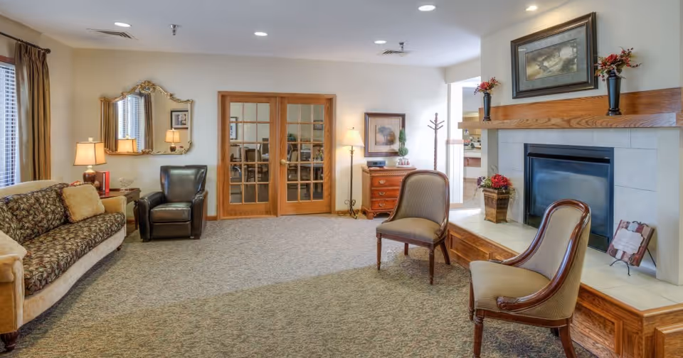 A cozy living room area with a patterned sofa, a dark leather armchair, two wooden chairs with cushioned seats, a fireplace with a wooden mantle decorated with flowers and a framed painting above it, a wooden side table with a lamp, and double glass doors leading to another room.