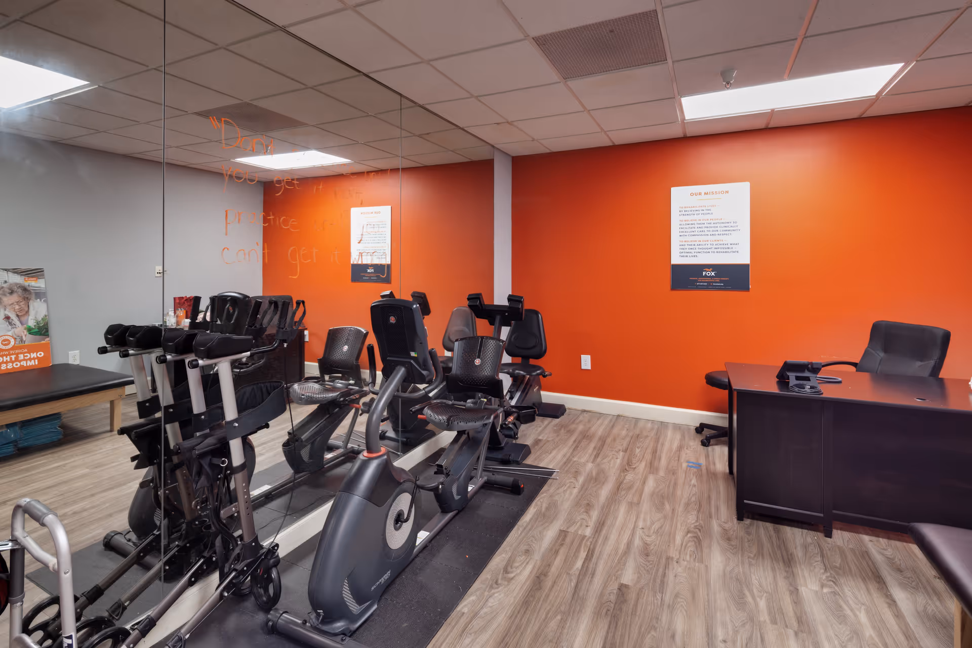 A fitness room with exercise equipment including stationary bikes and a treadmill, a large mirror on one wall, an orange accent wall with a framed mission statement, a black desk with a chair, and wood-style flooring.