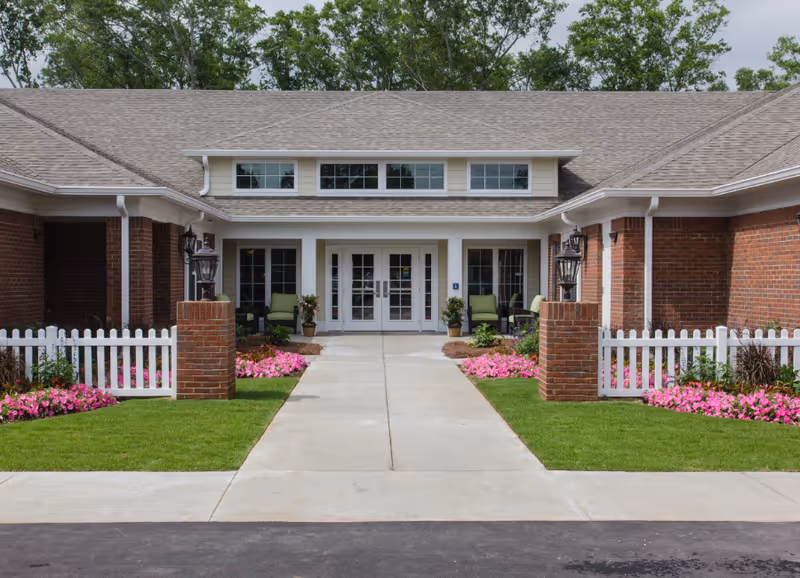 Front entrance of a senior living facility with a concrete walkway leading to double glass doors. The building has a brick exterior with white trim, a gray shingled roof, and white picket fences on either side of the walkway. There are green chairs and potted plants near the entrance, along with pink flowers lining the walkway.