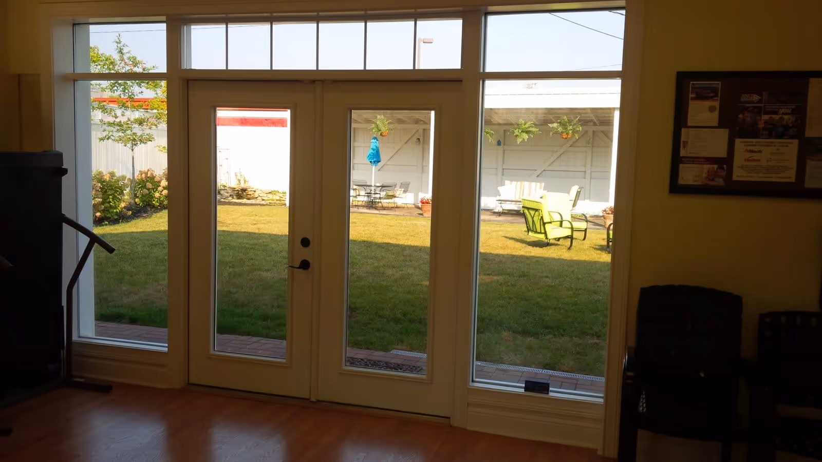 View through glass doors and windows from an interior room looking out onto a grassy outdoor patio area with chairs, tables, and hanging plants. Inside, there is a wooden floor and some chairs partially visible on the right side.