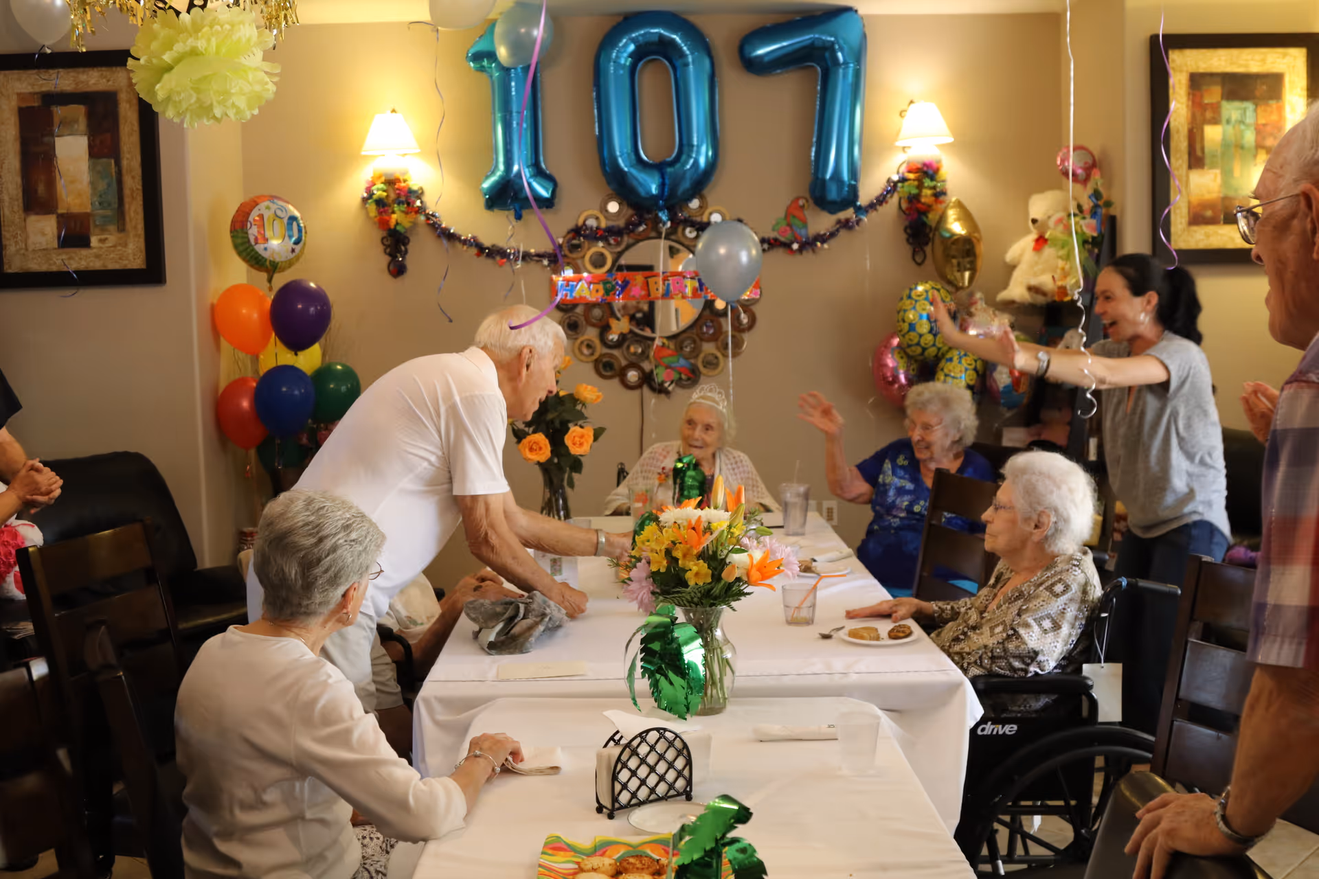 Seniors and caregivers gathered around a decorated dining table celebrating a 107th birthday with balloons and flowers.