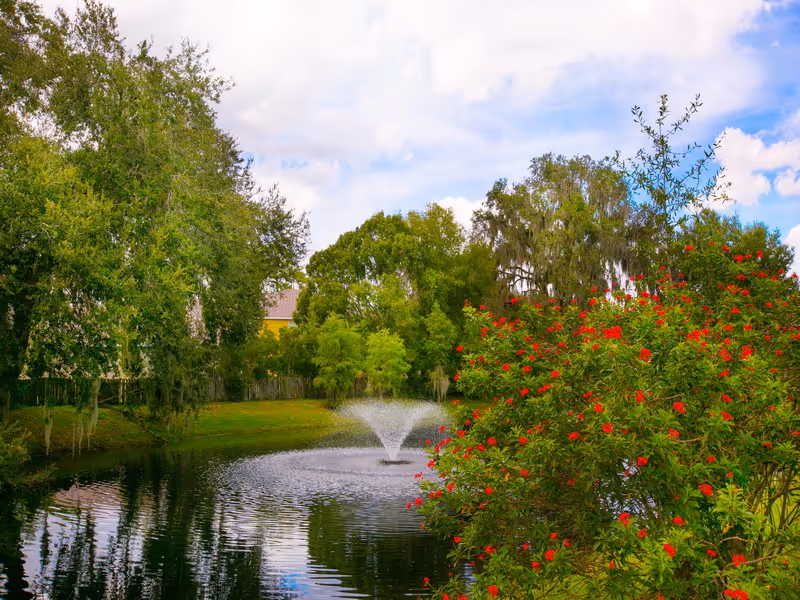 A peaceful outdoor scene featuring a pond with a water fountain in the center, surrounded by lush green trees and bushes. A bush with vibrant red flowers is visible on the right side, and the sky is partly cloudy with patches of blue.