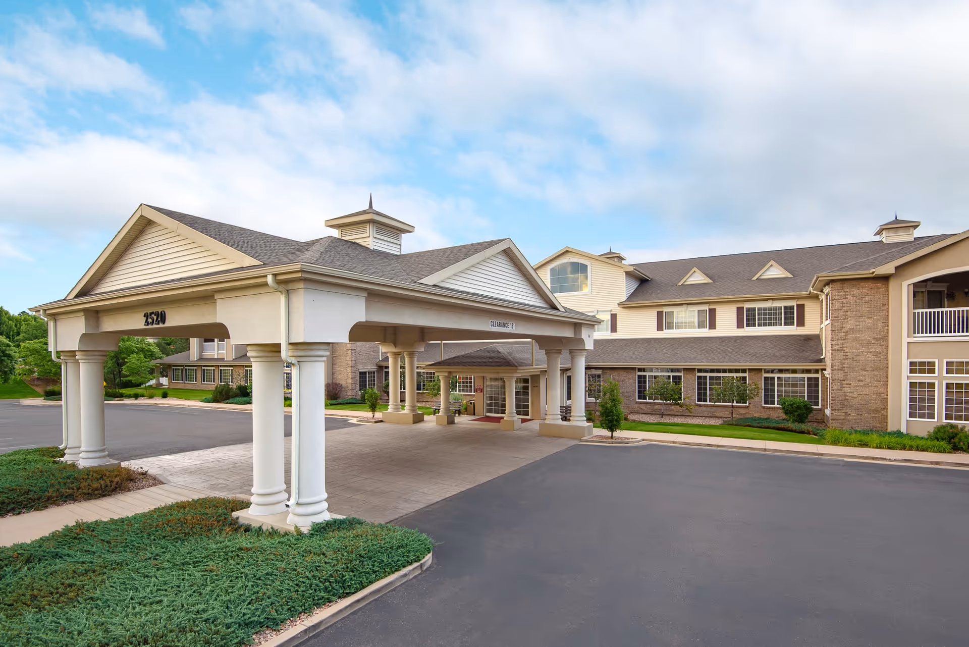 Exterior view of the Inn at Garden Plaza building featuring a covered entrance with columns, manicured landscaping, and a clear blue sky with some clouds.