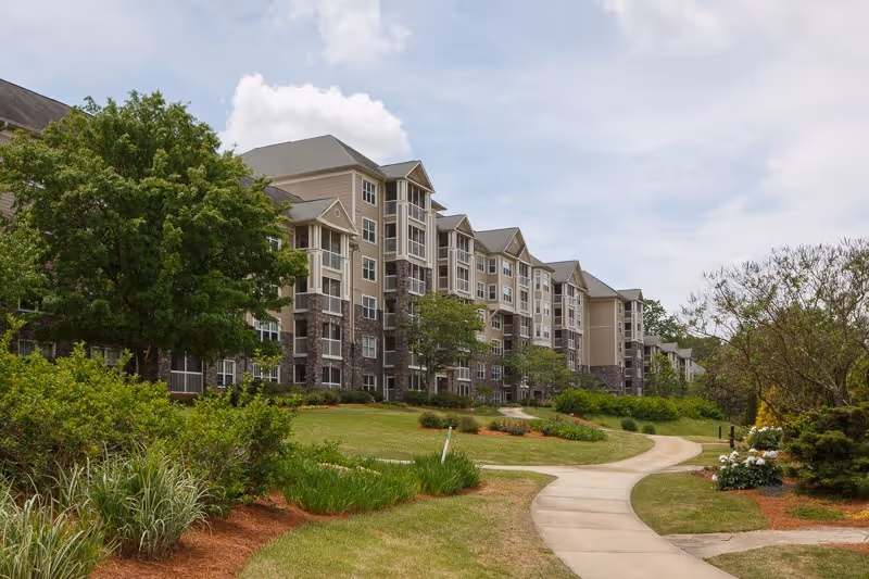 Exterior view of a multi-story senior living facility building with beige and stone facade, surrounded by green trees, shrubs, and a curved concrete walkway under a partly cloudy sky.