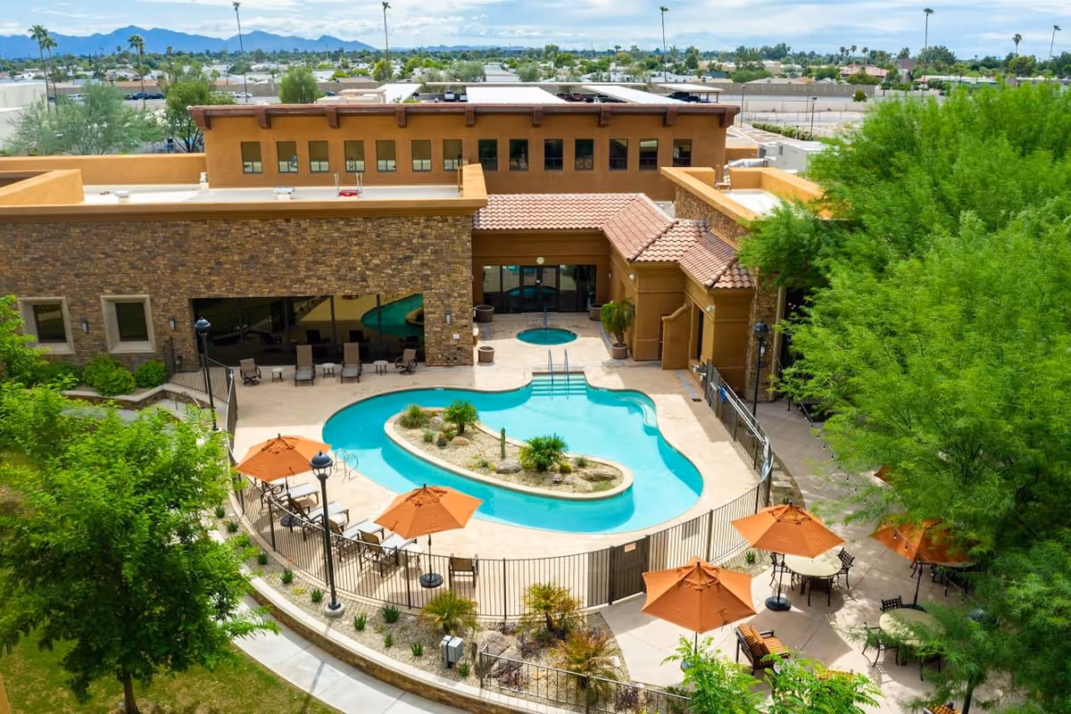 Aerial view of a courtyard with a kidney-shaped swimming pool, patio tables with orange umbrellas, and the surrounding senior living building.