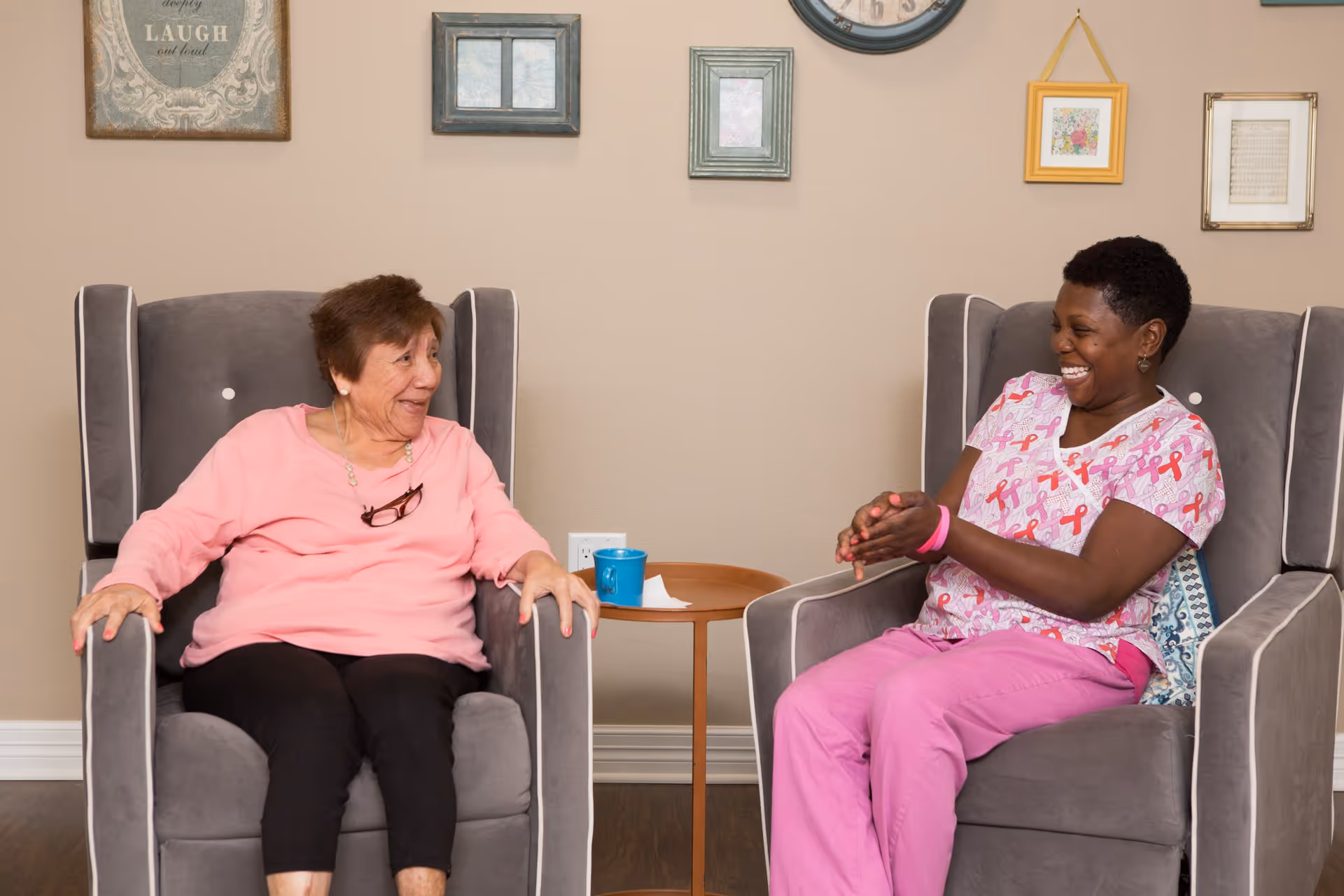 An elderly woman and a caregiver sitting in gray armchairs facing each other, smiling and engaging in conversation in a cozy room with framed pictures and a clock on the beige wall behind them.