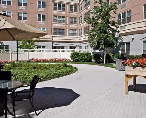 Outdoor courtyard area of a senior living facility with a paved walkway, green bushes, trees, and flower beds. There is a table with chairs and a large umbrella providing shade. The multi-story building surrounds the courtyard.