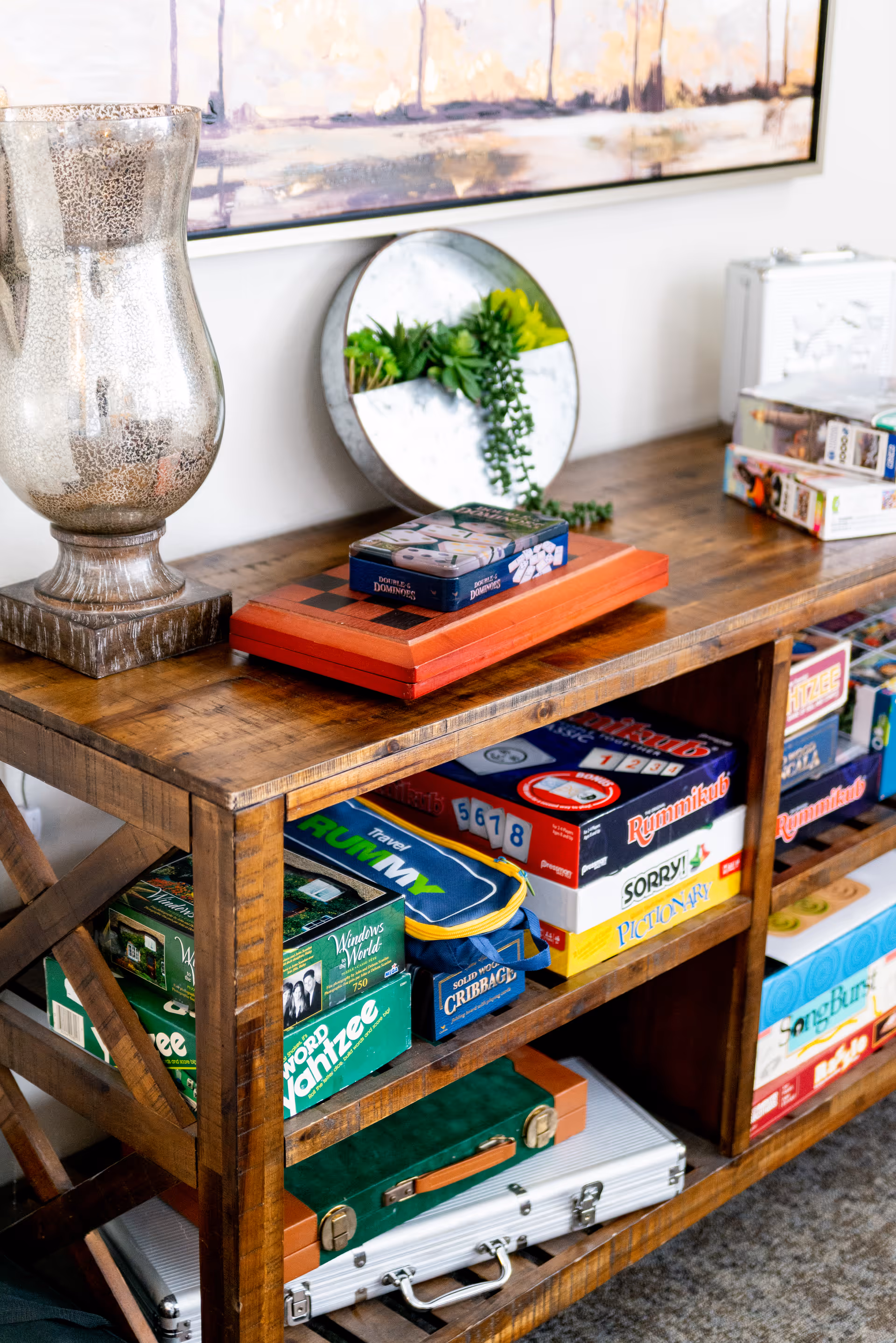 A wooden shelving unit filled with various board games including Rummikub, Sorry!, Pictionary, Travel Rummy, Word Yahtzee, and Cribbage. On top of the shelf is a decorative silver vase, a round mirror with green plants, and a wooden chess/checkers board with a box of dominoes on it. A framed abstract painting hangs on the wall above the shelf.