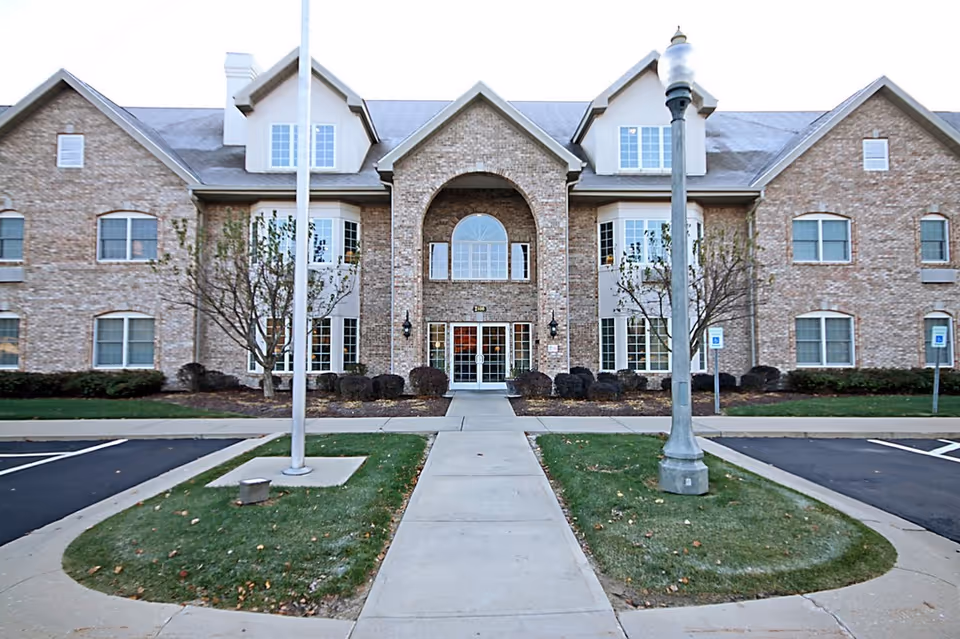 Front exterior view of a two-story brick building with multiple windows, a central entrance with glass doors, a sidewalk leading up to the entrance, two flagpoles, a streetlamp, and parking spaces on either side.