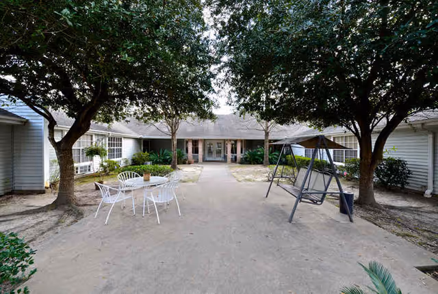 Outdoor courtyard area at Lucas Court featuring a concrete pathway, two large trees, a white metal table with four chairs, and a gray swing bench. The courtyard is surrounded by single-story buildings with windows and doors, and some greenery including bushes and potted plants.