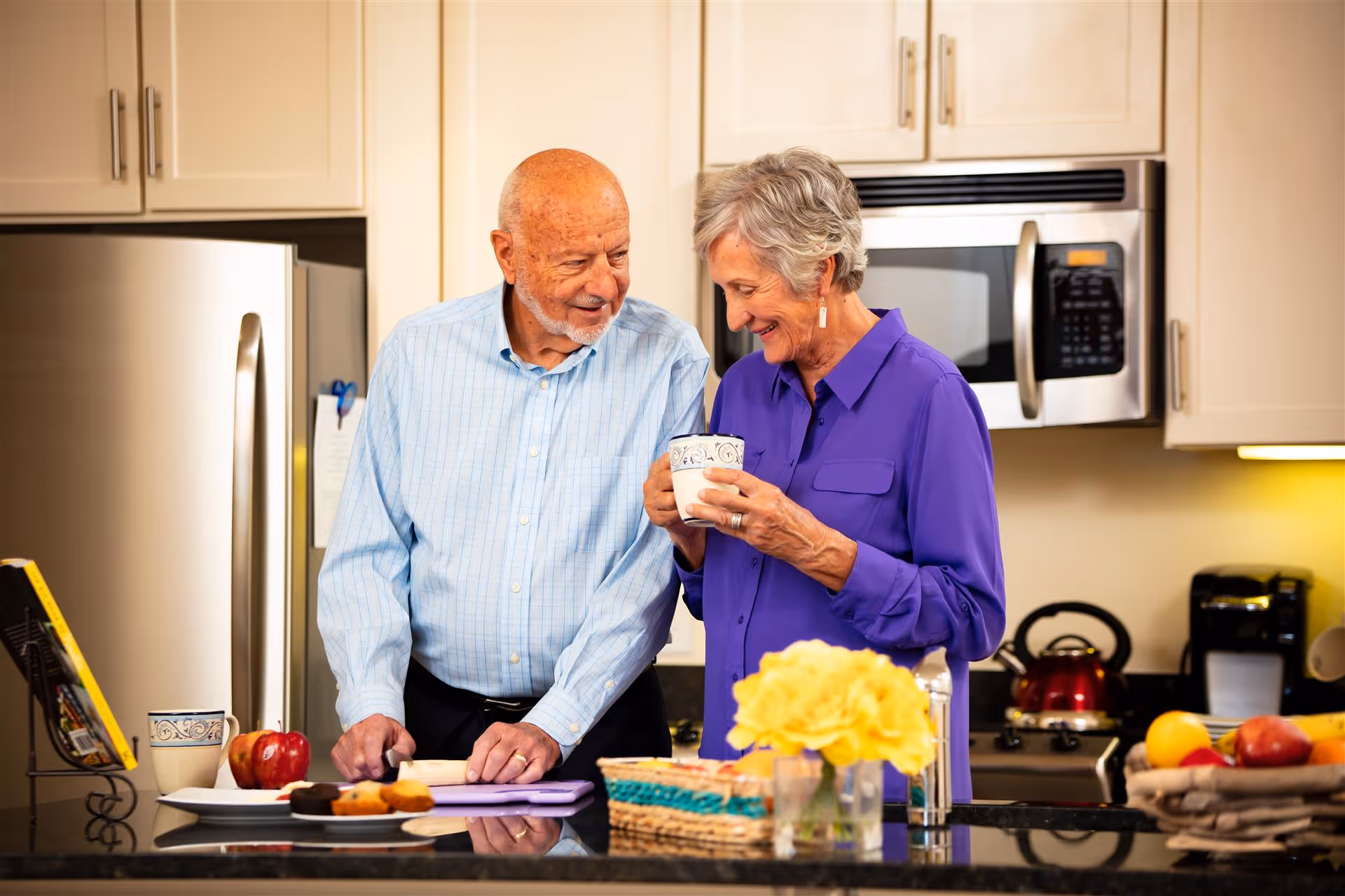 An elderly man and woman standing together in a kitchen. The man is wearing a light blue checkered shirt and is cutting something on a purple cutting board. The woman, dressed in a purple blouse, is holding a mug and smiling at the man. The kitchen features stainless steel appliances, including a refrigerator and microwave, with various items like fruit, flowers, and a coffee maker on the counter.