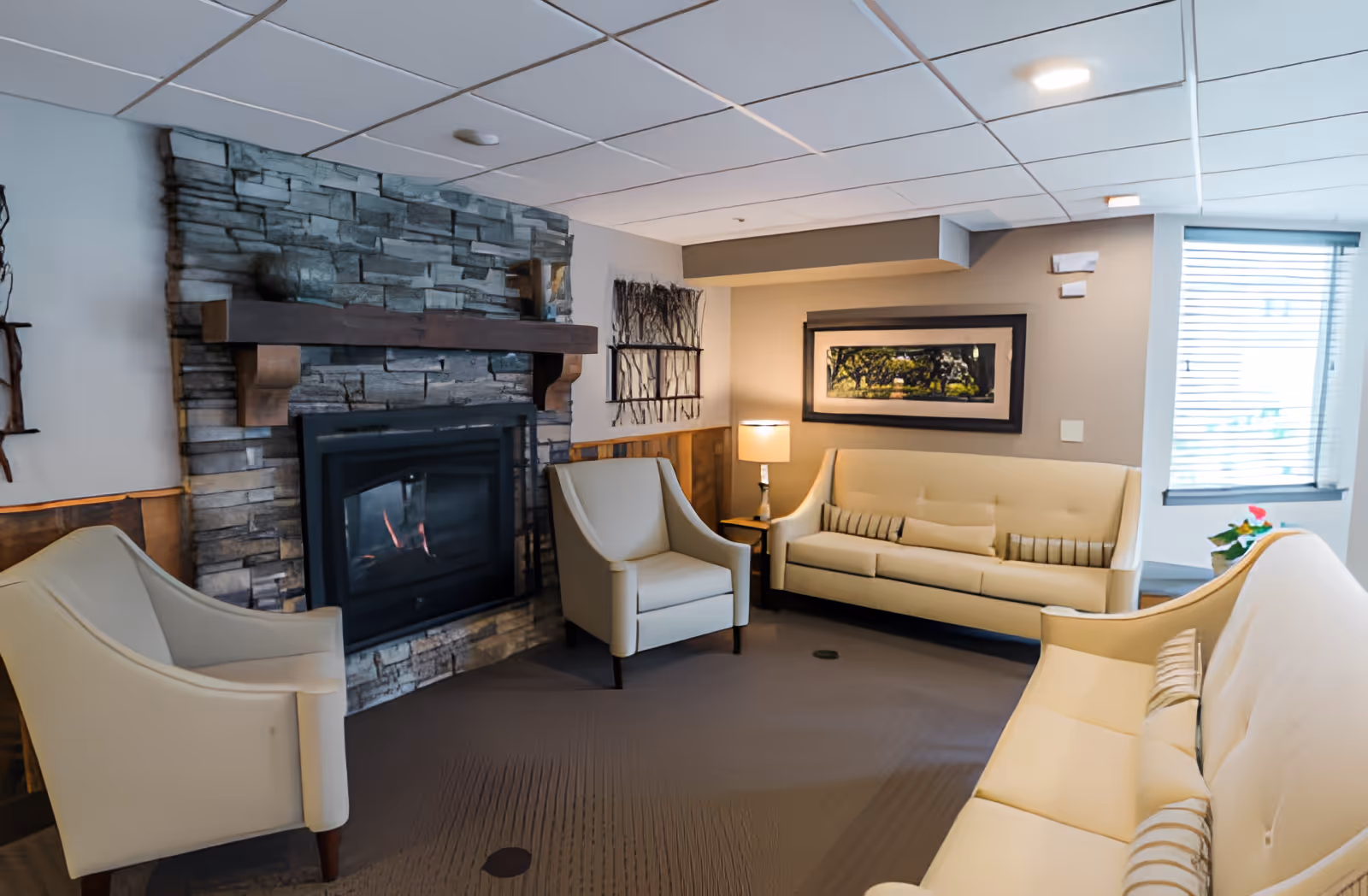 A cozy living room area with a stone fireplace, beige armchairs, and sofas arranged around a carpeted floor. There is a framed landscape picture on the wall, a table lamp on a small side table, and a window with blinds letting in natural light.