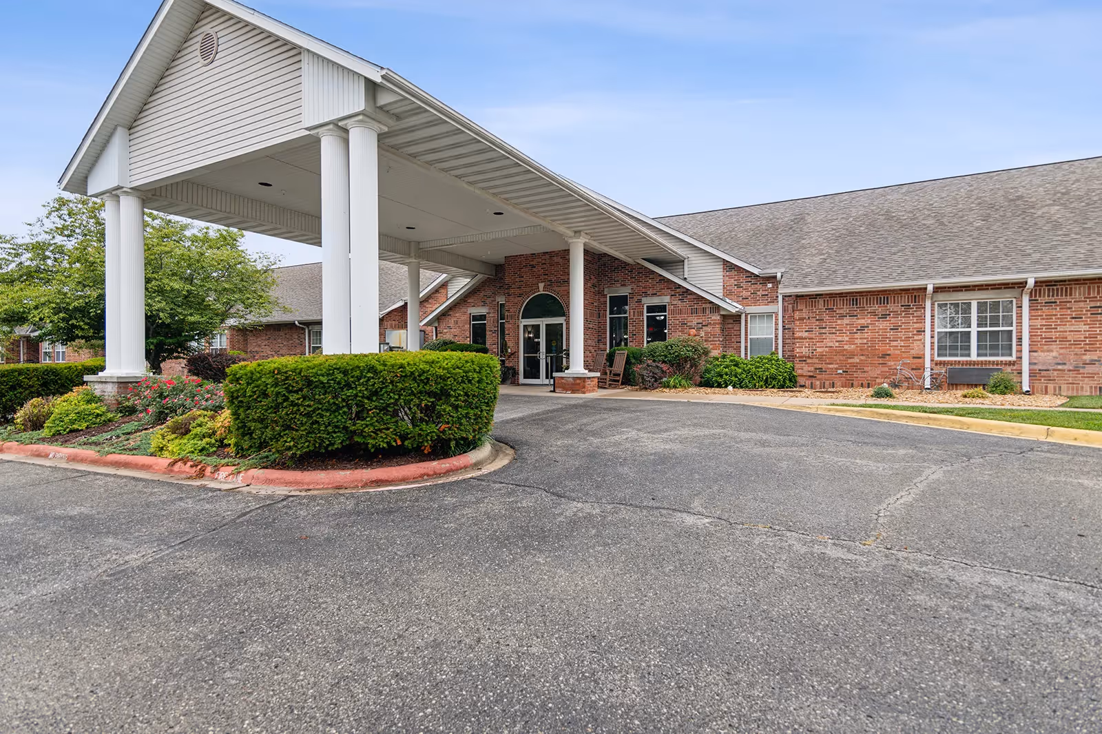 Front entrance of a single-story red brick building with a covered porte-cochère supported by white columns, landscaped shrubs, and a paved driveway.