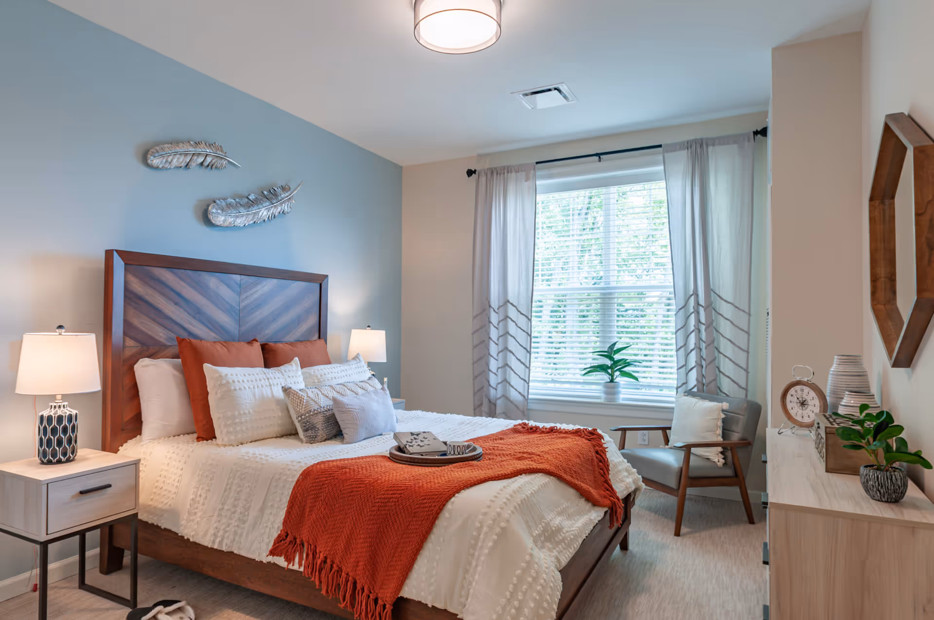 A cozy bedroom with a wooden bed frame and headboard, white textured bedding, and an orange throw blanket. There are multiple pillows on the bed. Two bedside tables each have a lamp. A window with white blinds and light curtains lets in natural light. A wooden dresser with decorative items and a chair with a cushion are also visible. The walls are painted in light blue and beige tones with two silver feather wall decorations above the bed.