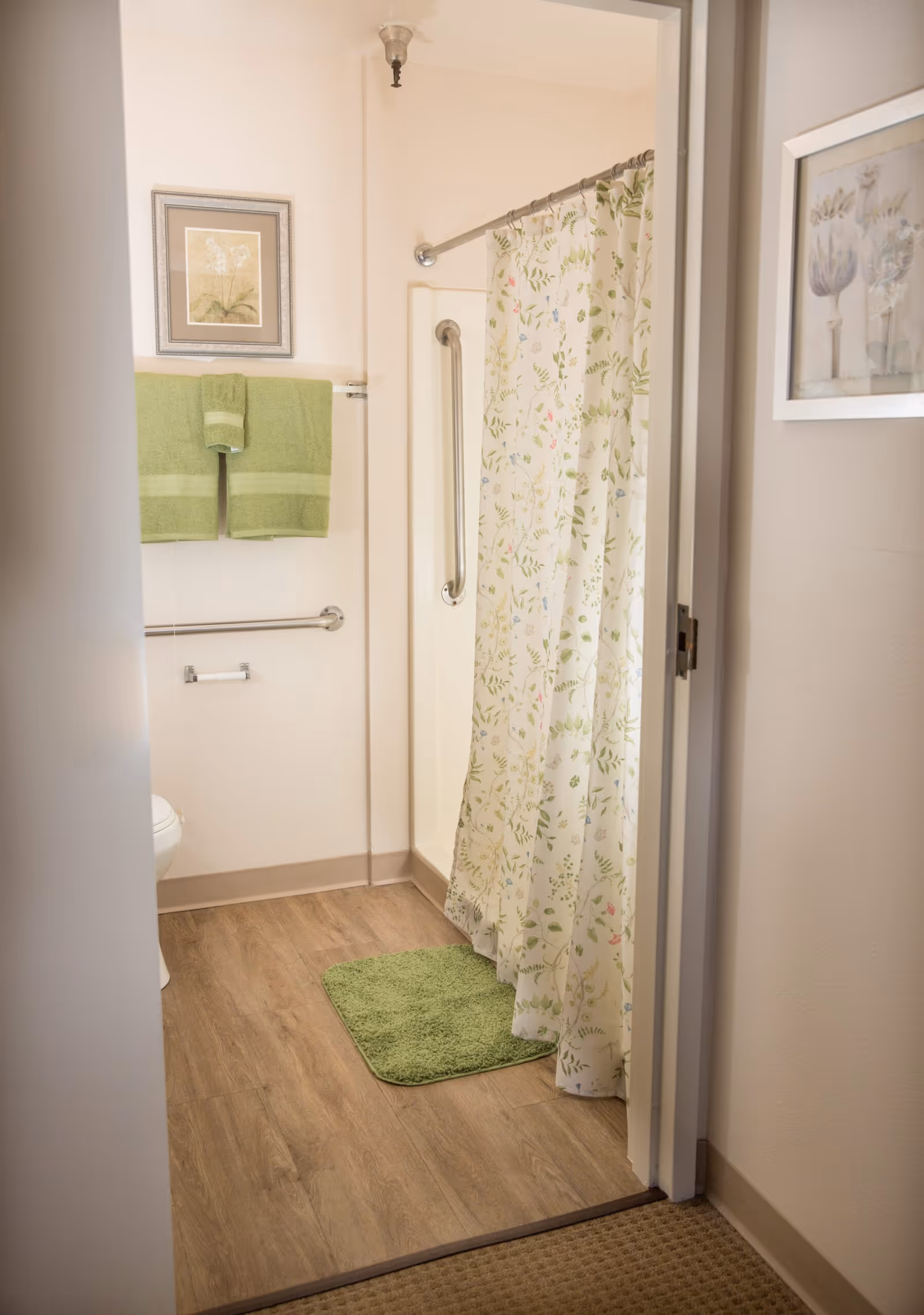 View into a bathroom with a floral shower curtain, green bath mat, and green towels hanging on a towel rack. The bathroom has wood-look flooring, grab bars near the shower and toilet, and framed botanical artwork on the walls.