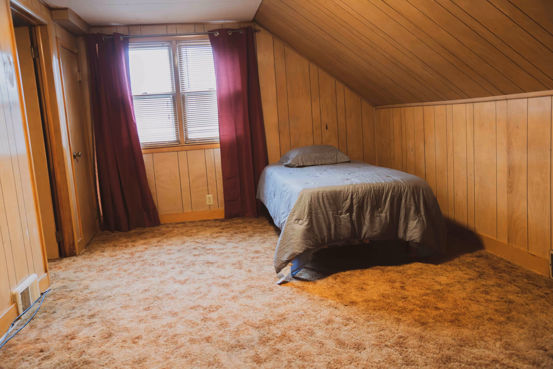 A small bedroom with wood-paneled walls and a slanted ceiling. There is a single bed with a gray comforter and pillow positioned against the right wall. A window with closed blinds and dark red curtains is on the back wall. The floor is covered with a brown carpet.