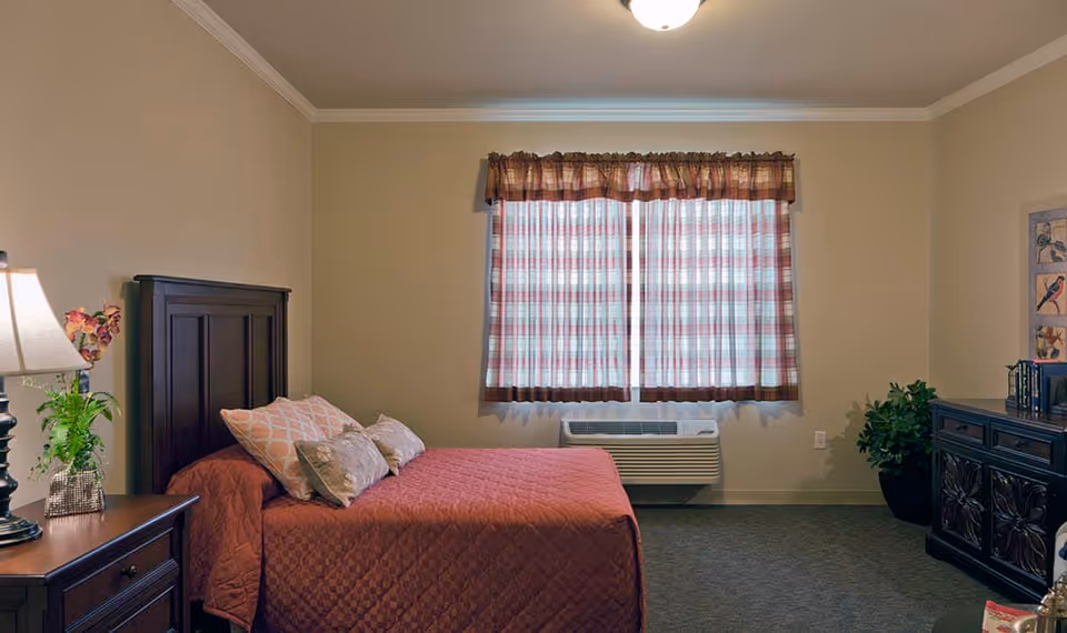 A cozy bedroom with a bed covered in a red quilt and several pillows. There is a wooden nightstand with a lamp and a small plant on it. A window with checkered curtains is centered on the back wall above an air conditioning unit. On the right side, there is a dark wooden dresser with decorative carvings and a potted plant next to it. The walls are painted beige and the carpet is dark gray.
