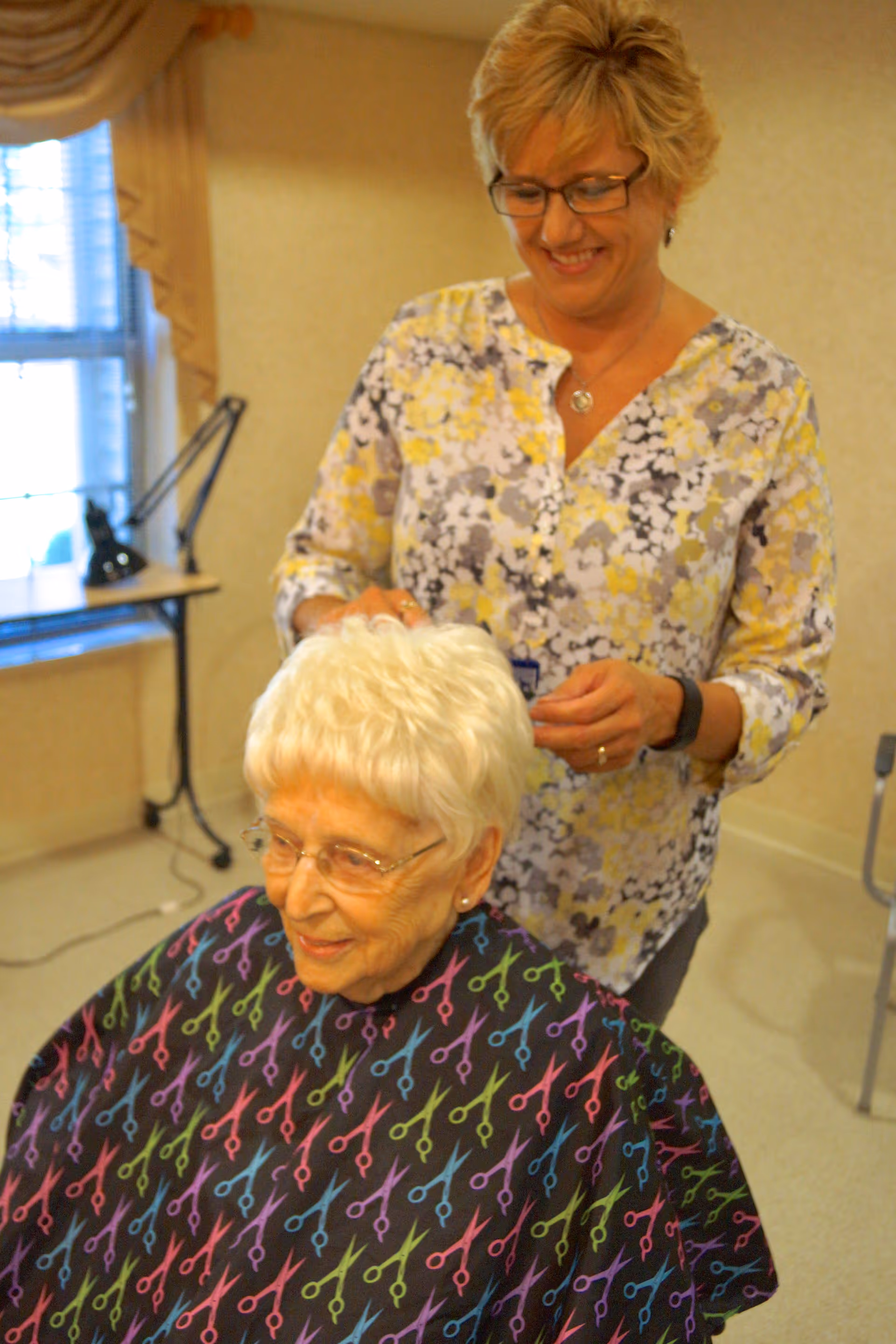 An elderly woman with white hair and glasses is seated wearing a colorful haircut cape with scissors pattern, while a smiling woman standing behind her styles her hair in a well-lit room with a window and a desk lamp in the background.