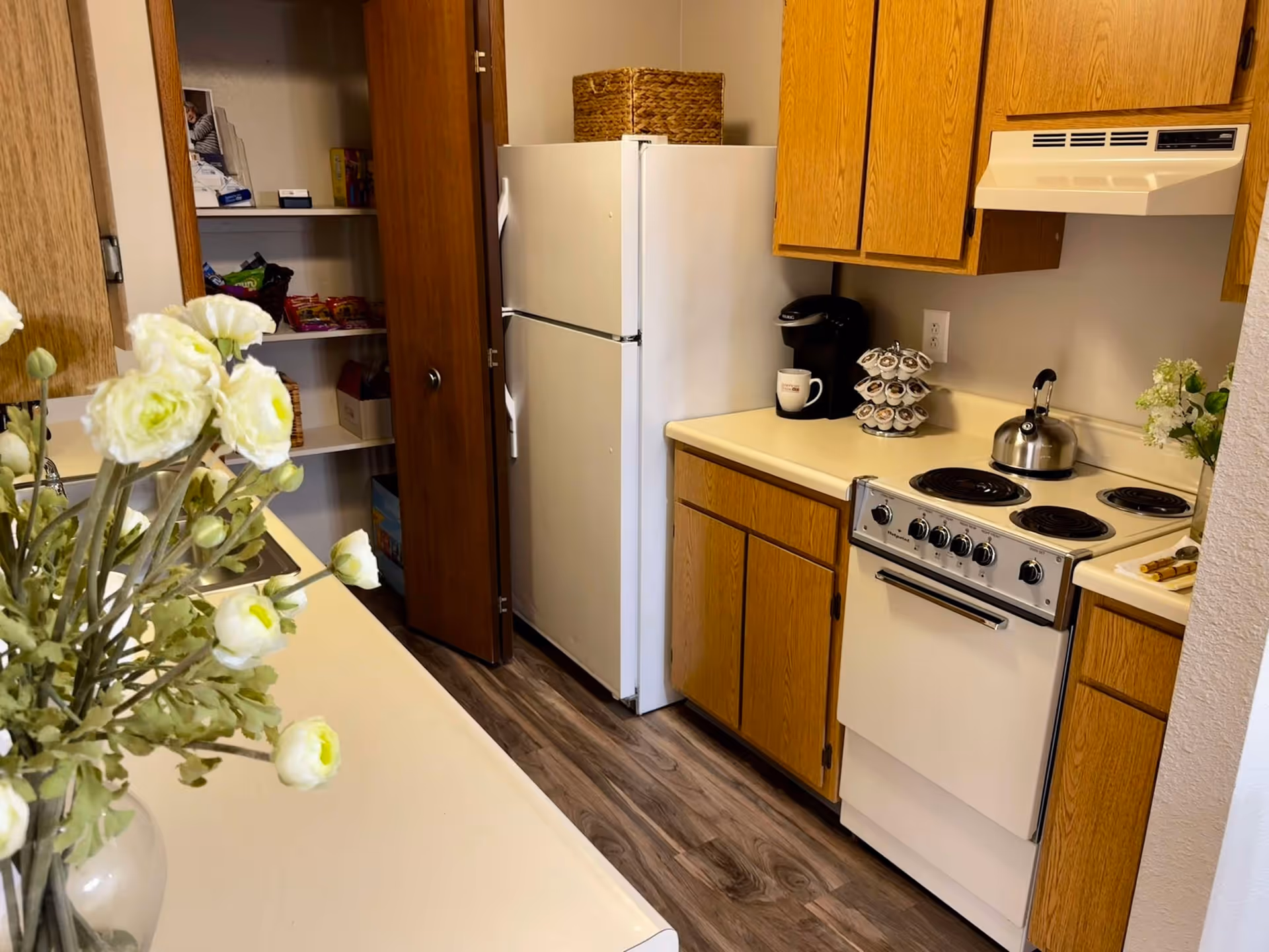 A small kitchen with wooden cabinets, a white refrigerator, an electric stove with four burners, a coffee maker, and a spice rack on the countertop. There is a pantry with open wooden doors showing shelves stocked with various food items. A vase with white flowers is placed on the counter in the foreground.