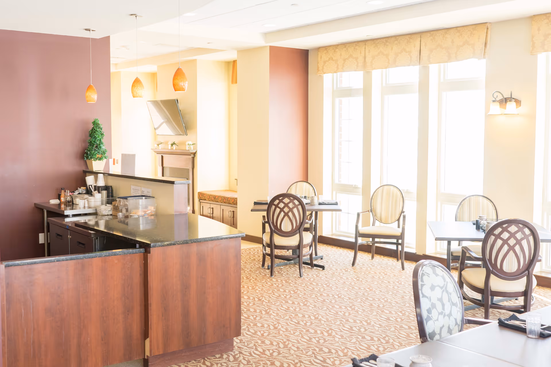 Bright dining area with several tables and chairs near large windows allowing natural light. A counter with a dark granite top and wooden base is on the left side, with hanging pendant lights above it. The room has a warm color palette with beige walls and patterned carpet.