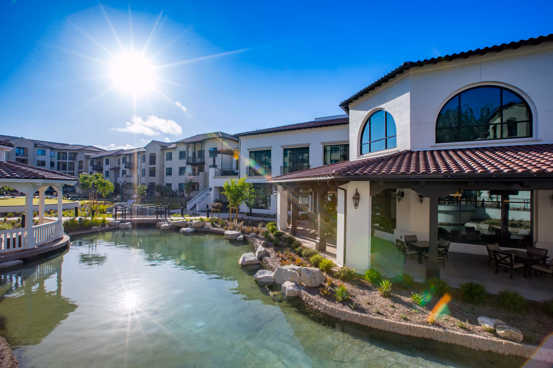 Sunny outdoor view of The Glen at Scripps Ranch senior living facility featuring a clear pond with rocks along the edge, a covered patio area with seating, and multi-story buildings with balconies in the background under a bright blue sky.