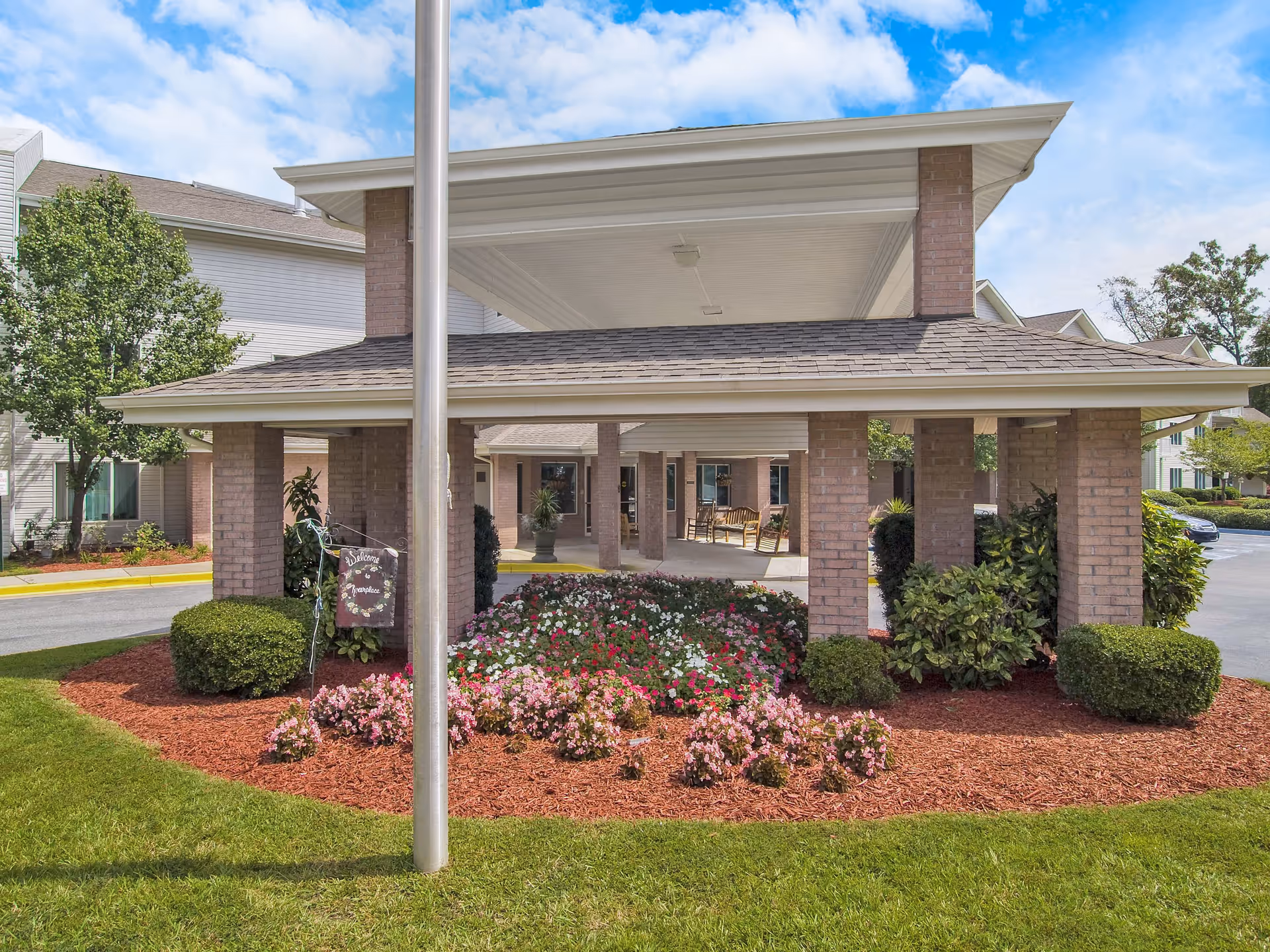 Covered entrance area of a senior living facility with brick pillars, a flower bed with various colorful flowers, neatly trimmed bushes, and a flagpole in the foreground. The building and parking area are visible in the background under a partly cloudy blue sky.