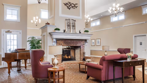 Bright communal living room with a central brick fireplace, chandeliers, a piano, and upholstered seating arranged around tables.