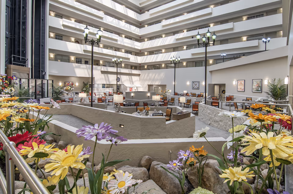 A spacious indoor atrium area in a senior living community featuring multiple tables and chairs arranged for dining or socializing. The space is surrounded by several floors with balconies and railings, illuminated by natural light from above. Decorative streetlamp-style lights and framed artwork adorn the walls. In the foreground, colorful artificial flowers and rocks add a garden-like touch.