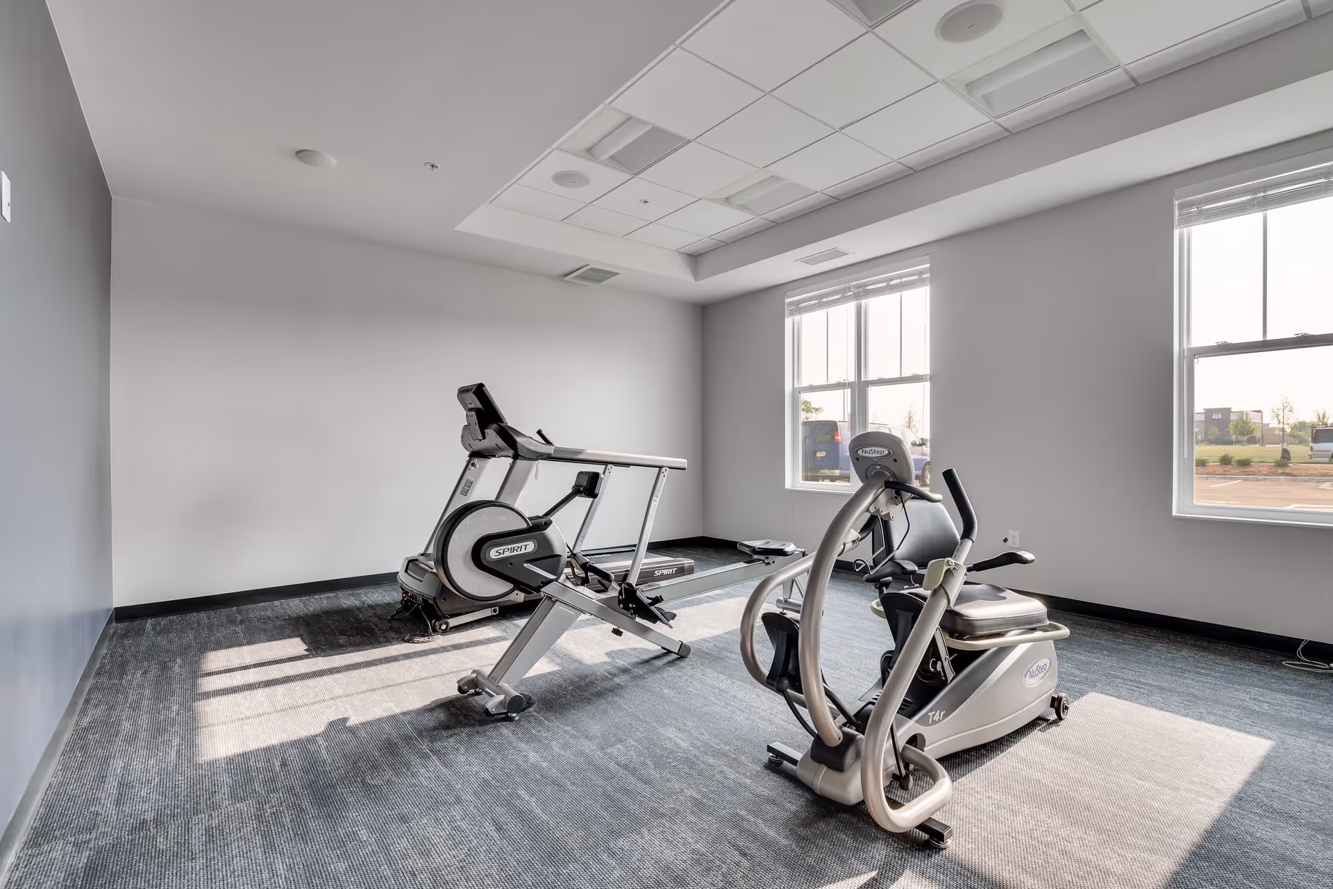 A small sunlit fitness room with two cardio machines (a recumbent bike and an elliptical) on a carpeted floor and two windows.