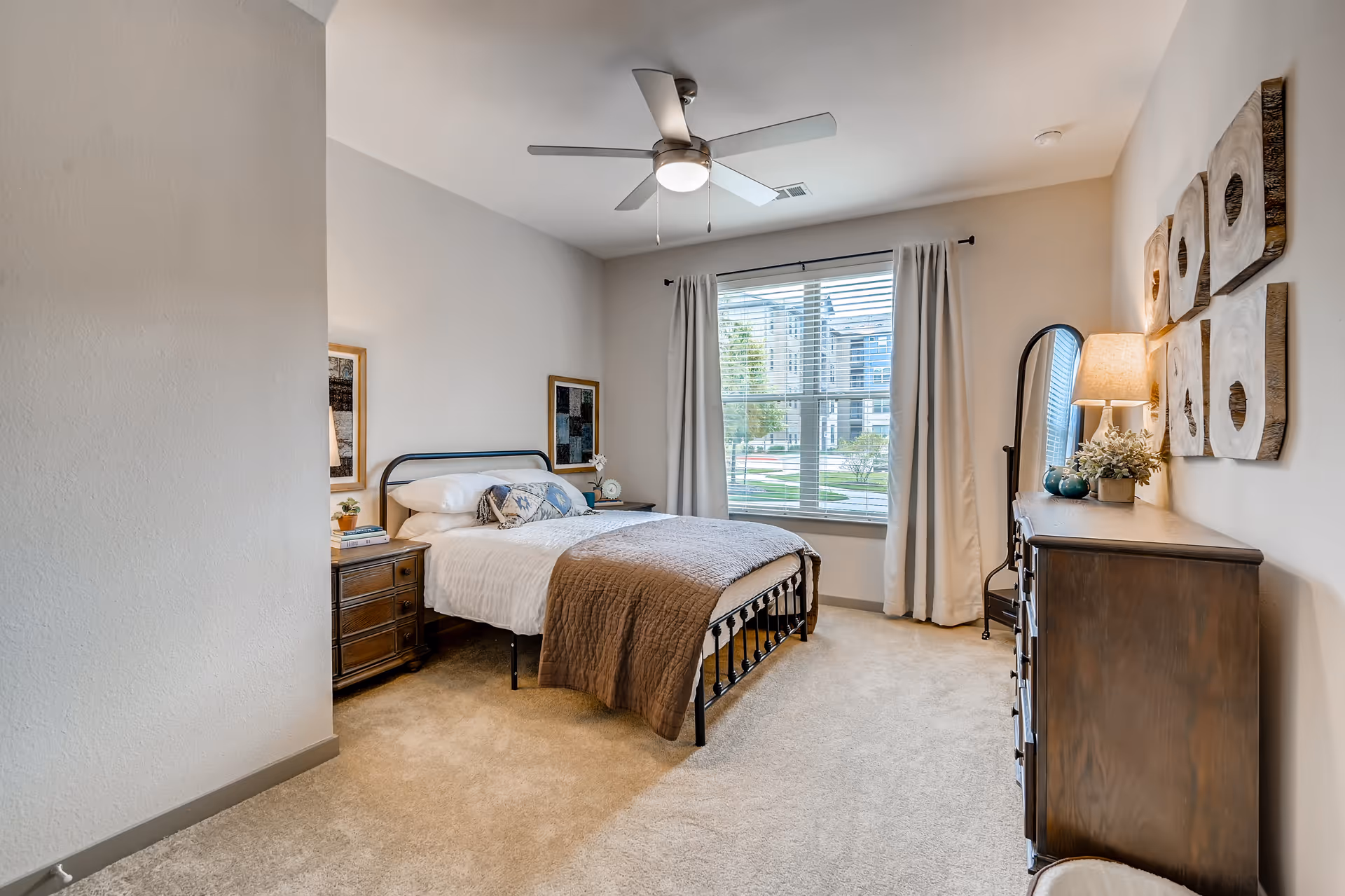 Bright furnished bedroom with a metal-framed bed, nightstand, dresser, window with curtains, ceiling fan, and neutral decor.