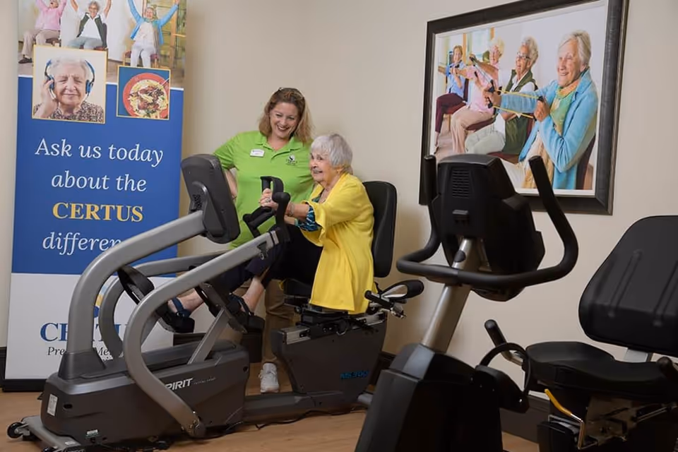 An elderly woman in a yellow cardigan is seated on a recumbent exercise bike, smiling while a staff member in a green shirt stands beside her, offering support. The room contains exercise equipment and a banner that reads 'Ask us today about the CERTUS difference.' A framed picture of elderly women exercising is on the wall behind them.