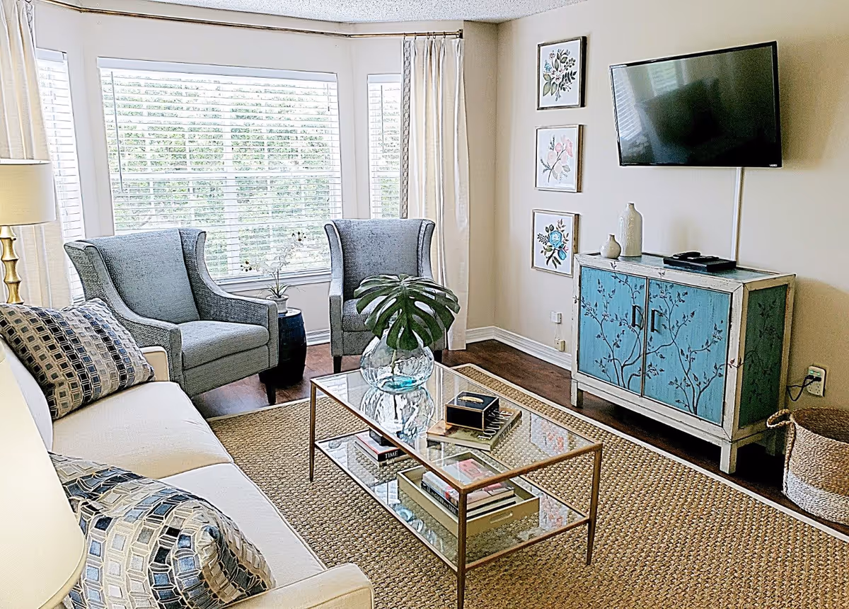 A bright and cozy living room with a large window covered by white blinds and curtains. The room features a white sofa with patterned cushions, two gray armchairs, a glass coffee table with books and a vase holding a large green leaf, a blue cabinet with floral designs, three framed floral artworks on the wall, a flat-screen TV mounted above the cabinet, and a woven basket on the floor.
