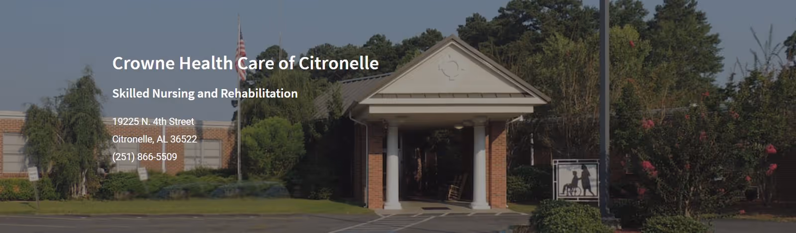 Exterior front view of Crowne Health Care of Citronelle, a skilled nursing and rehabilitation facility with a covered entrance supported by white columns, surrounded by trees and bushes under a clear sky.