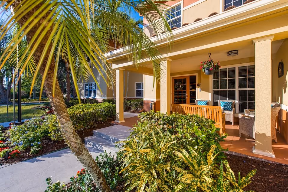 Outdoor view of a senior living facility porch area with a wooden bench, wicker chairs with blue patterned cushions, hanging flower pot, and lush green plants and palm trees surrounding the walkway leading to the entrance.