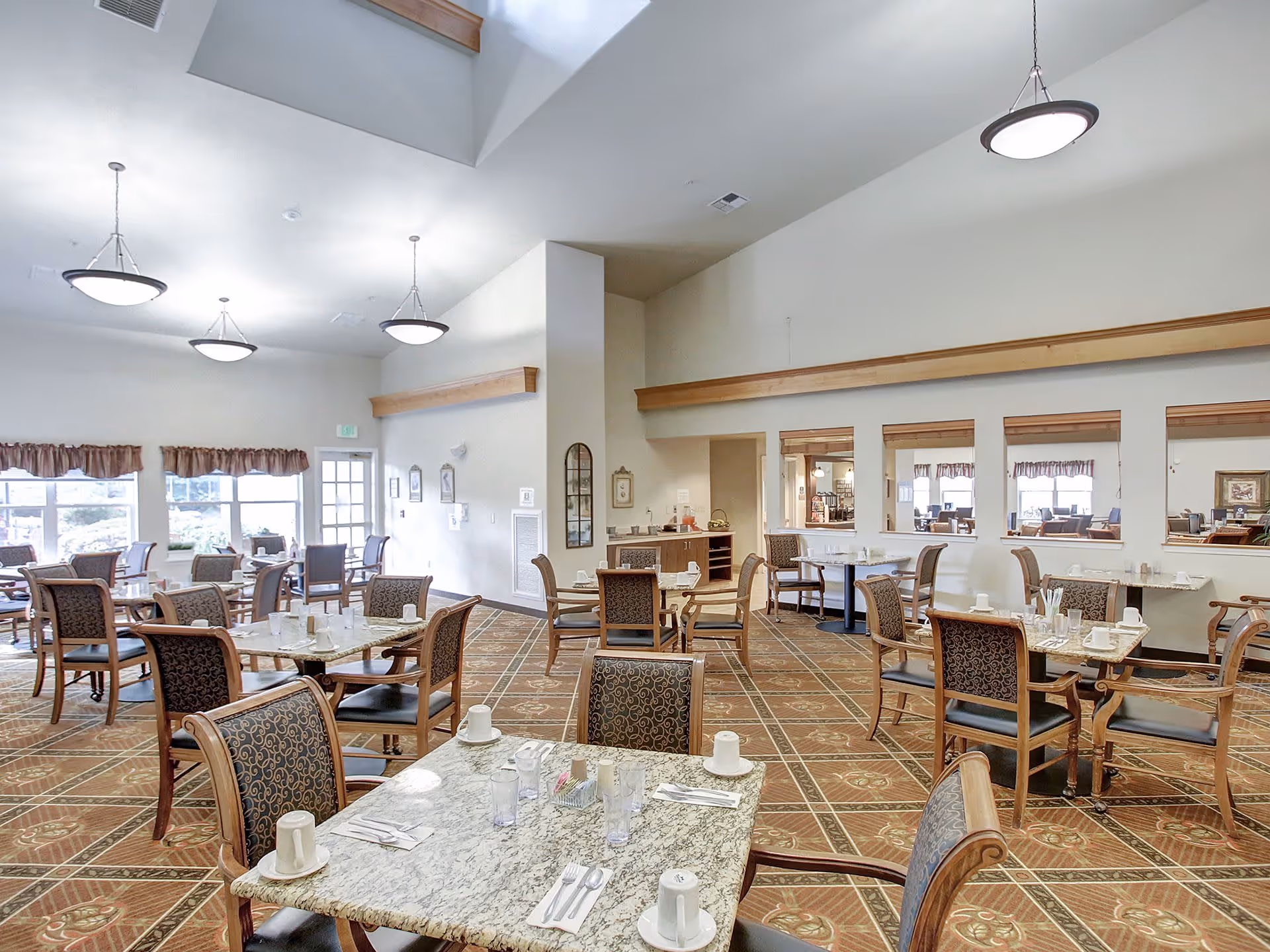 A spacious dining room in a senior living facility with multiple tables and chairs arranged neatly. Each table is set with cups, glasses, and utensils. The room has large windows with valance curtains allowing natural light to fill the space. The ceiling is high with hanging light fixtures, and the floor is covered with patterned carpet.