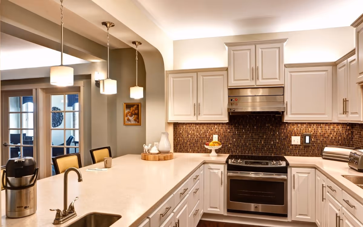 Modern kitchen with white cabinetry, a stainless steel oven and range hood, a mosaic tile backsplash, and a large countertop island with a sink and faucet. Three pendant lights hang above the island, and there are double glass doors leading to another room in the background.