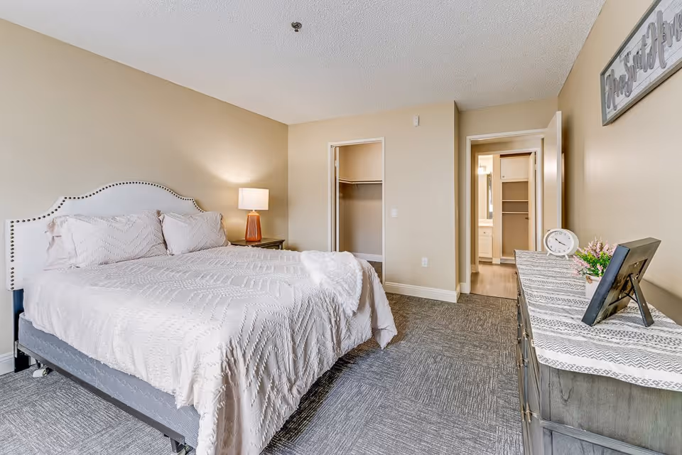 A neatly made bed with a white headboard and light-colored bedding in a senior living bedroom. There is a bedside table with a lamp on the left side of the bed. On the right side of the room, there is a dresser with a decorative runner, a clock, a small plant, and a framed photo. The room has beige walls and carpeted flooring. Two open doorways are visible, one leading to a closet and the other to a bathroom area.