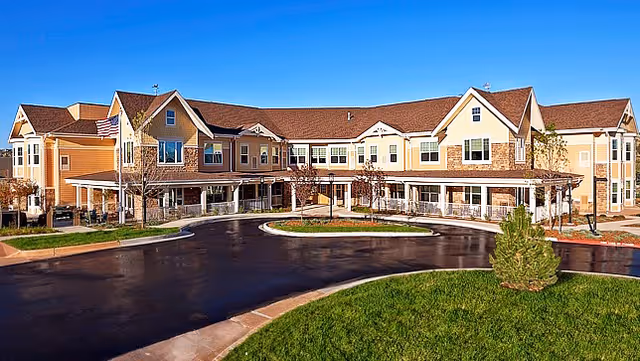 Front exterior of a two-story assisted living building with a curved driveway and landscaped lawn under a clear blue sky.